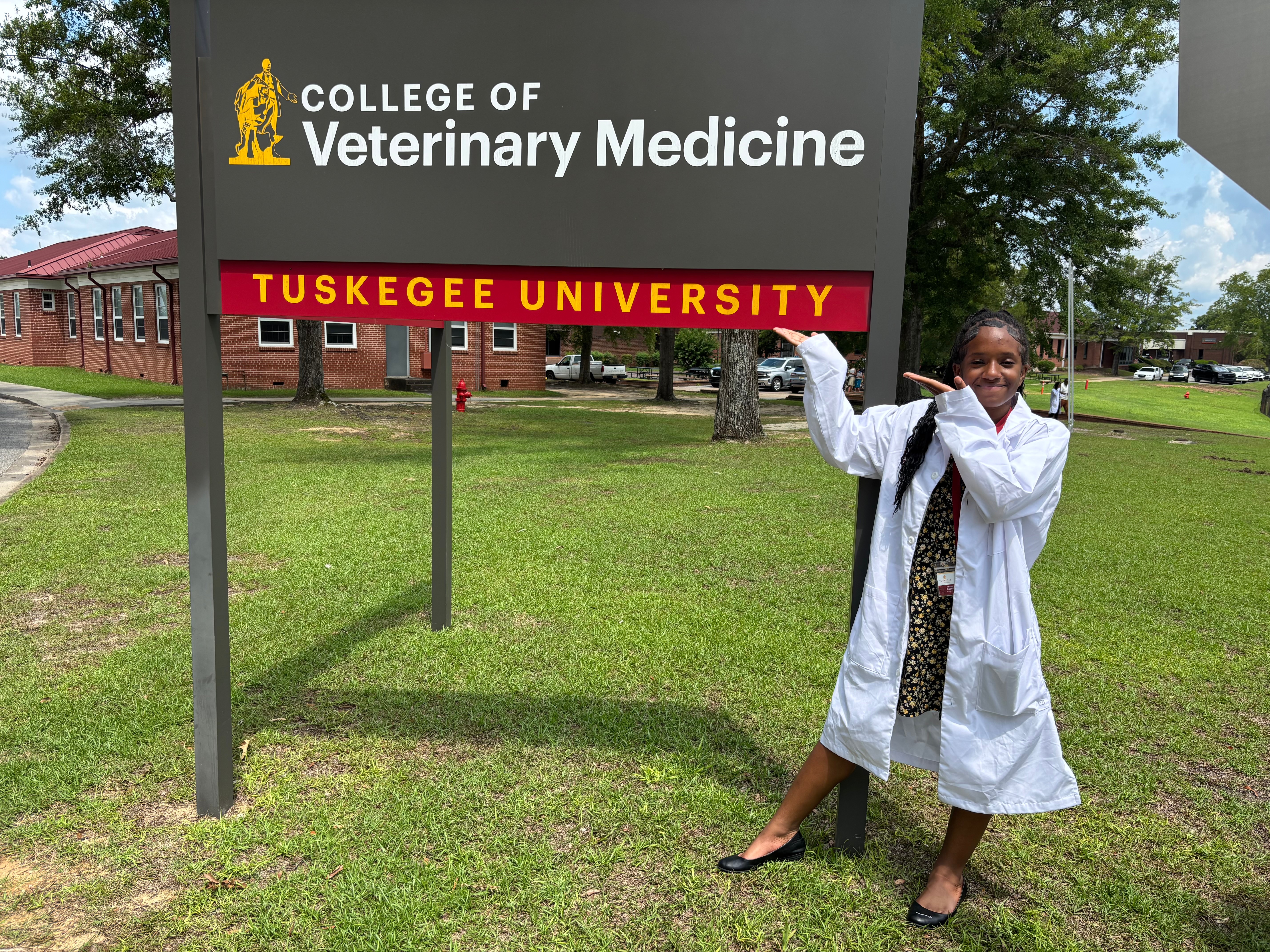 Child in a white coat pointing at a sign for College of Veterinary Medicine and Tuskegee University outdoors.