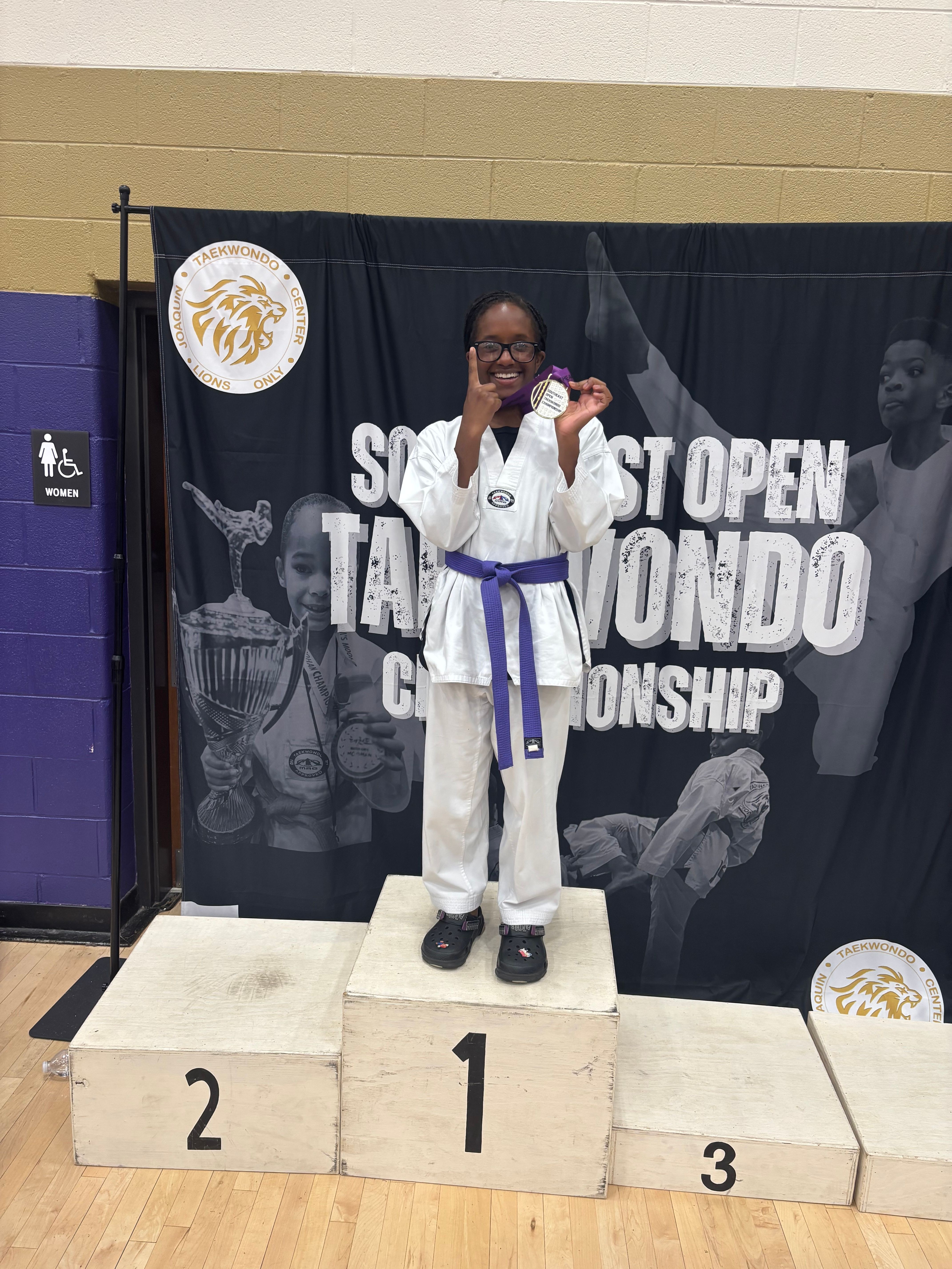 Person in a white martial arts uniform with a purple belt standing on a winners' podium, holding a trophy, with a black backdrop and a medal around their neck.