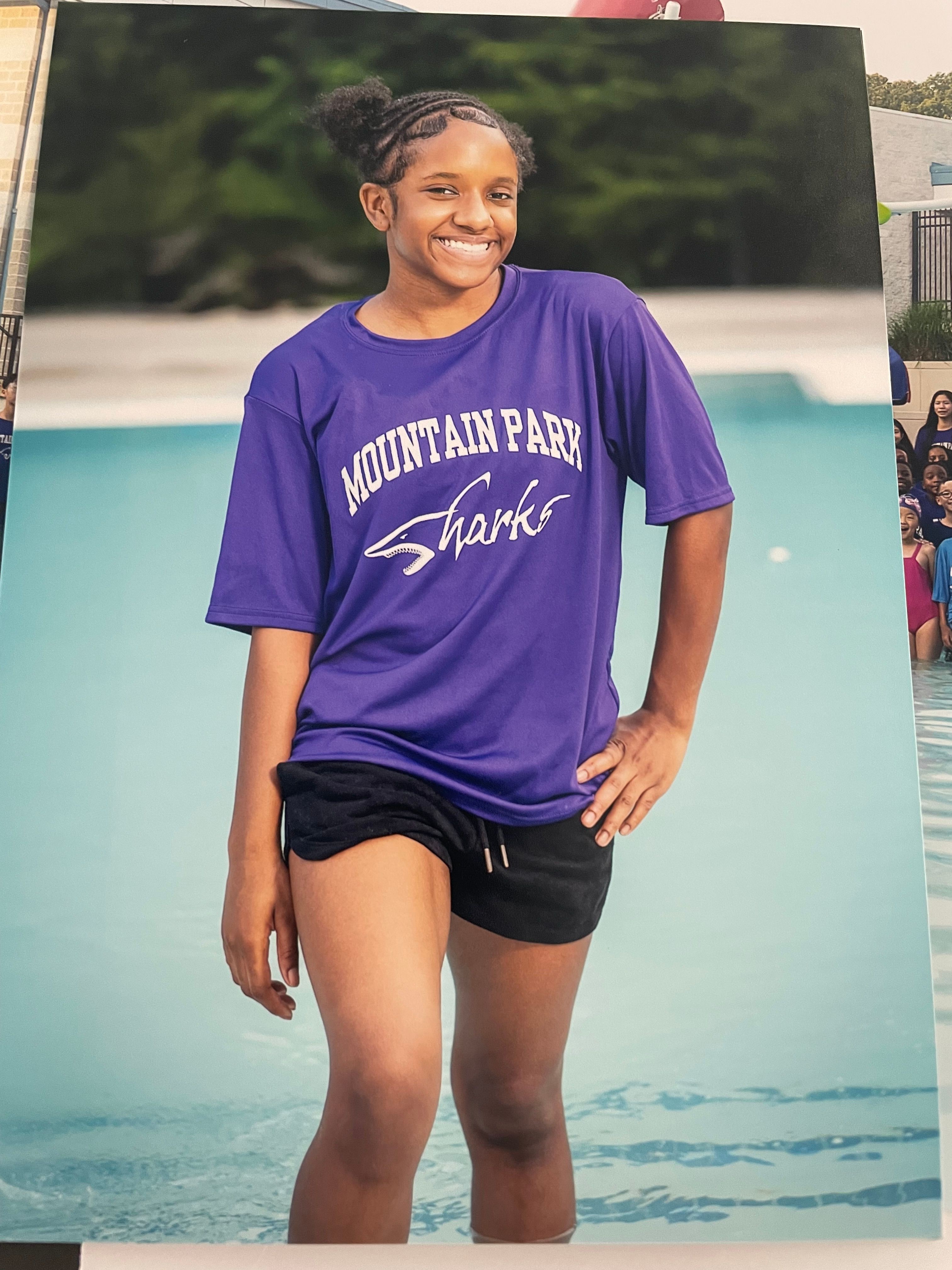 Person standing on a poolside with water in the background, wearing a purple T-shirt and black shorts.
