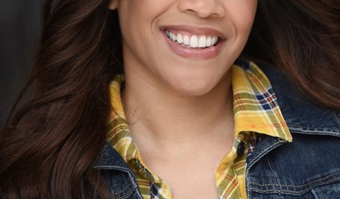 Young woman with long dark hair smiling, wearing a yellow plaid shirt and denim jacket, outdoors.