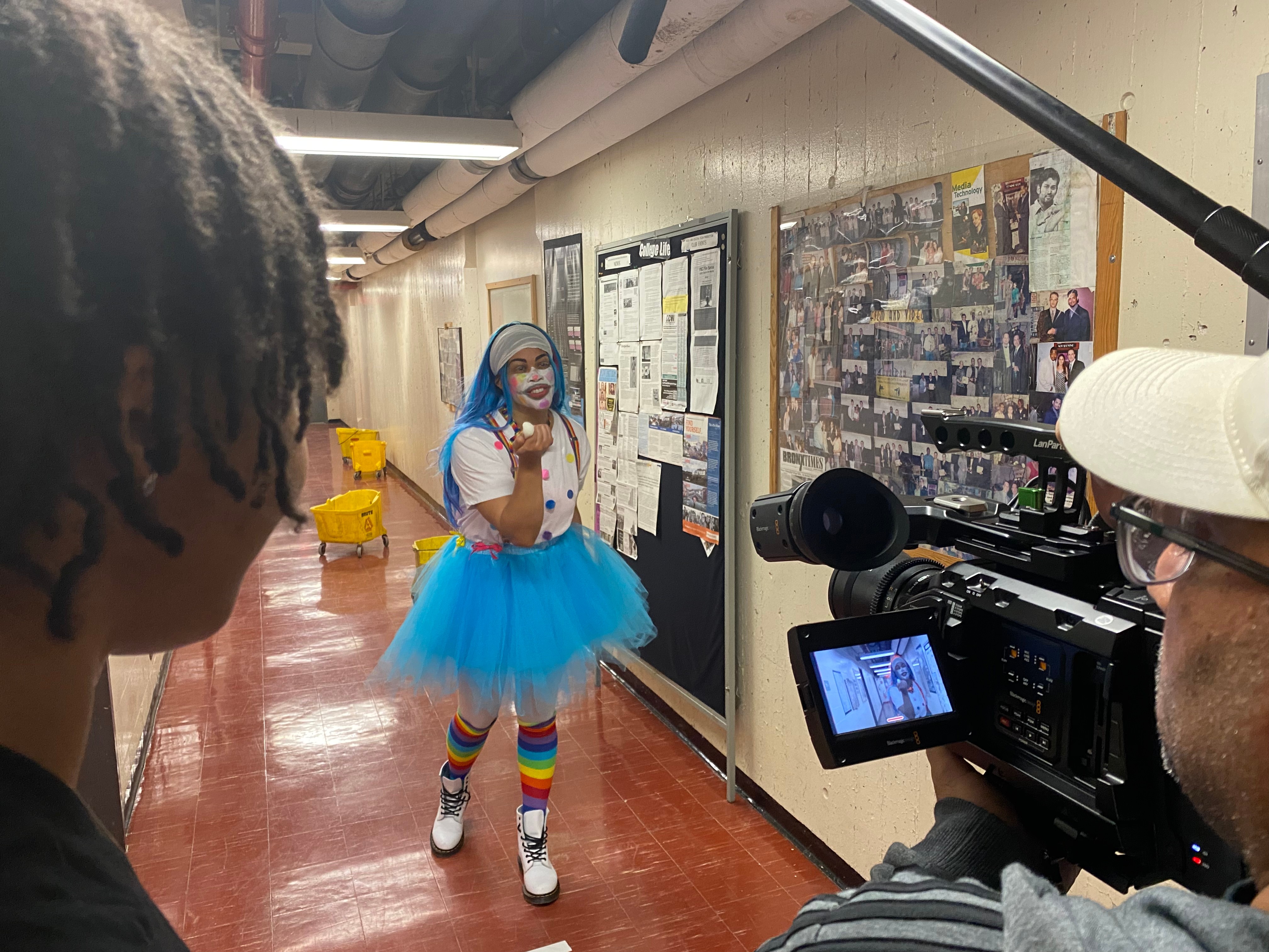 Person dressed as a clown in a blue tutu and rainbow socks being filmed in a hallway with bulletin boards.