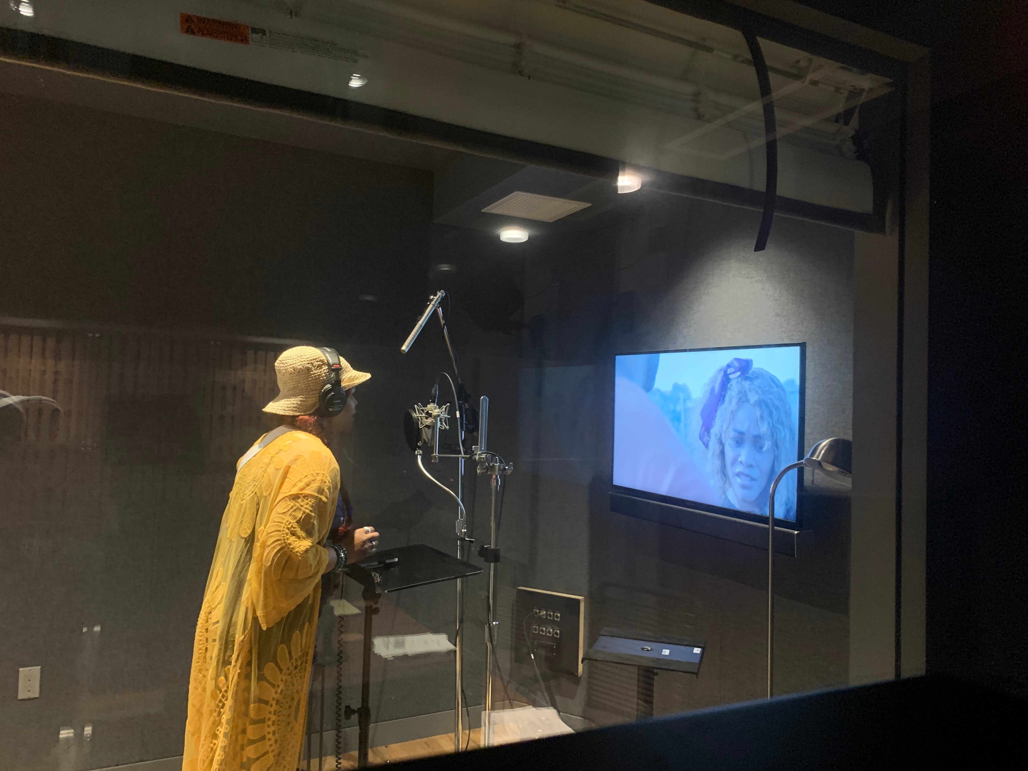 Person in yellow coat and hat looking at a monitor in a recording studio, with equipment and a TV screen showing a face.