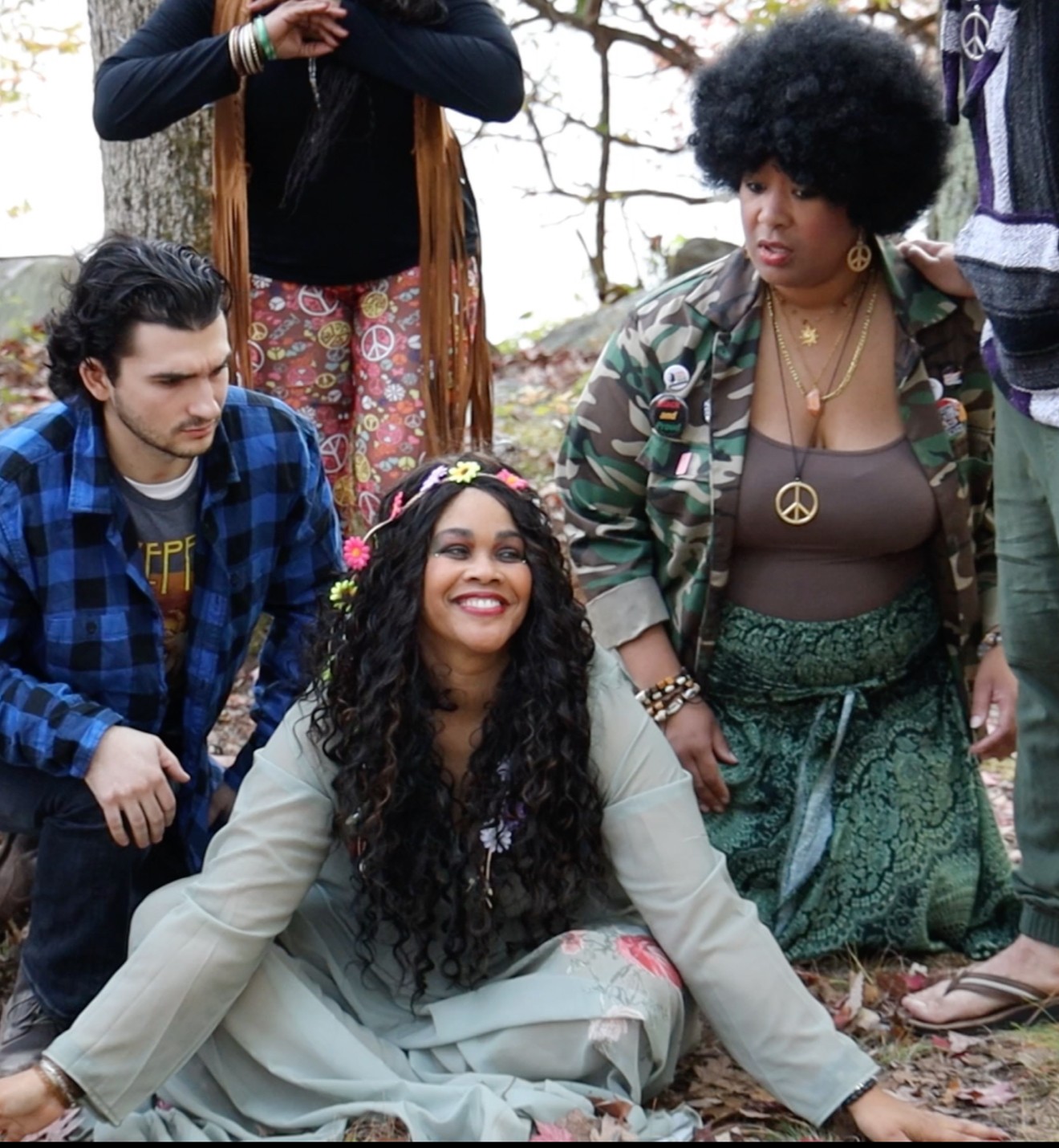 Group of diverse people outdoors, woman sitting on ground smiling, others standing around, trees in background.