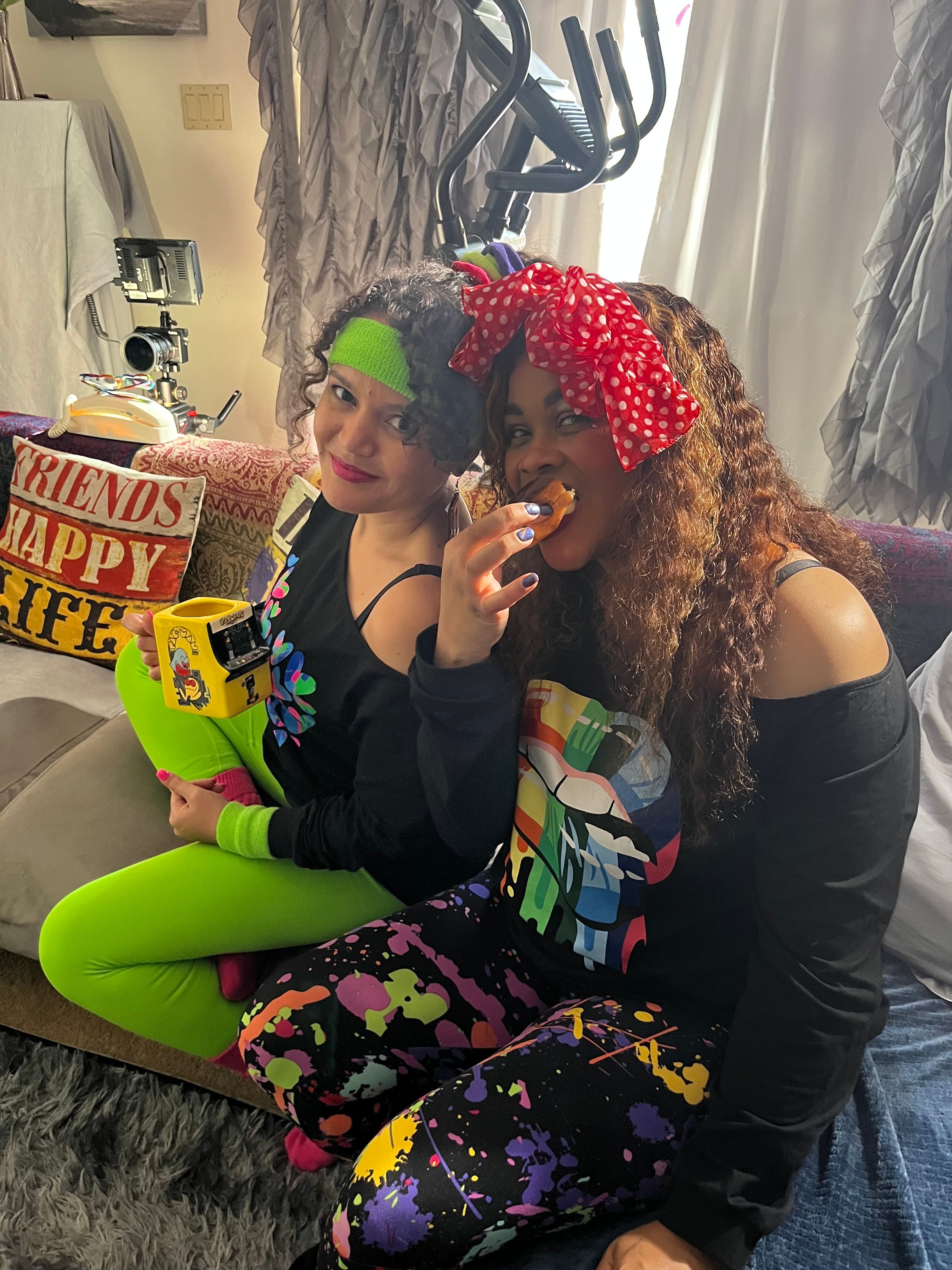 Two women sitting on a couch, one with curly hair and a red polka dot headband, the other with a green headband, smiling and holding a snack. The woman with curly hair is wearing a black top and bright green pants. The woman with the green headband is wearing a black top with colorful patterns. A box of Fritos and a yellow bag are on the couch. Background shows a window with curtains and a table with items.