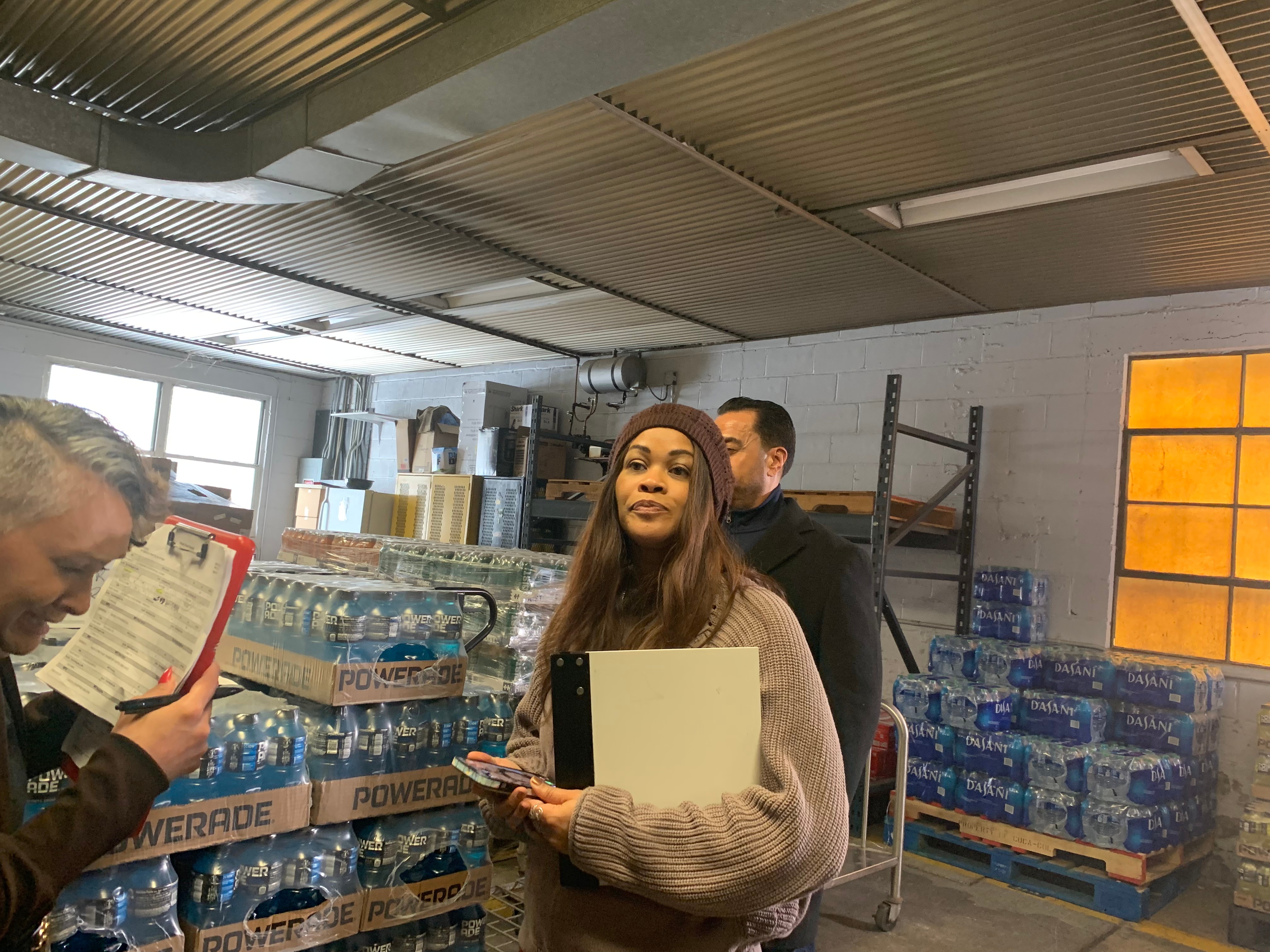 Group of people in a warehouse with water bottles, one woman holding a folder and phone, others reading documents.