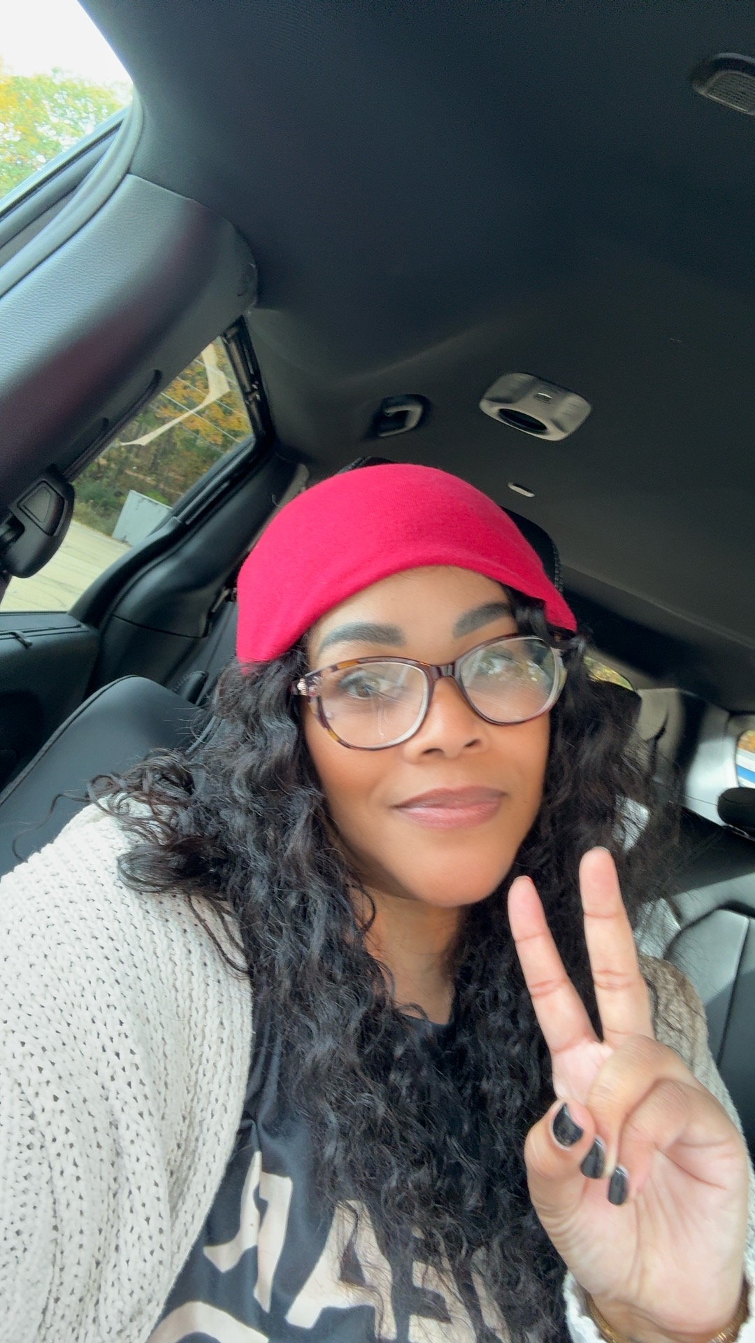 Woman with glasses and curly hair wearing a red cap inside a car, making a peace sign with her hand.