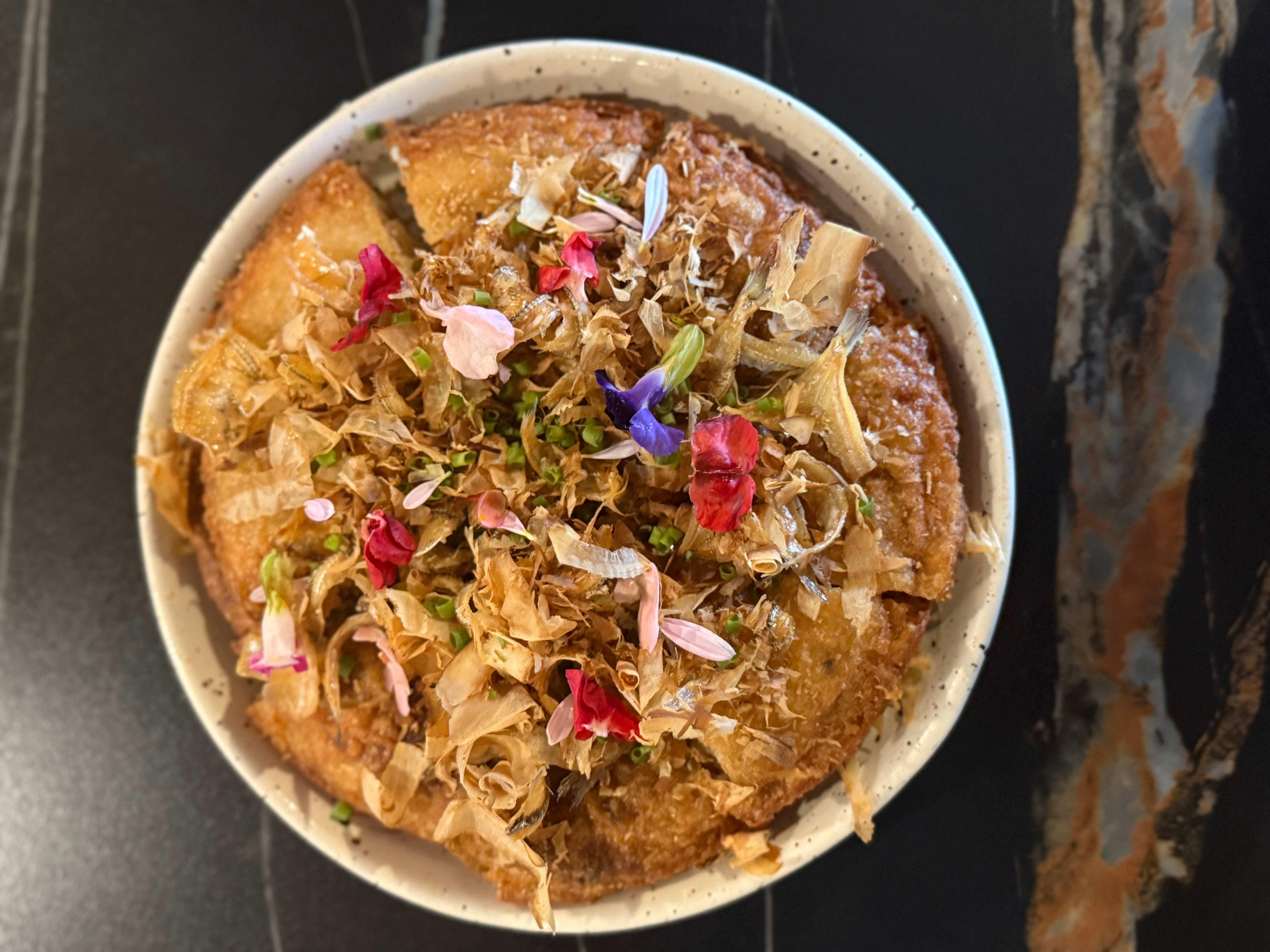 Bowl of dessert topped with colorful edible flowers and shredded toppings, served on a dark surface.