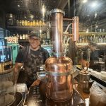 Person standing behind a copper distillation apparatus in a bar setting with bottles and glasses in the background.