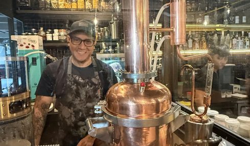 Person standing behind a copper distillation apparatus in a bar setting with bottles and glasses in the background.