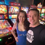 A smiling man and woman take a selfie in front of pinball machines at an arcade.