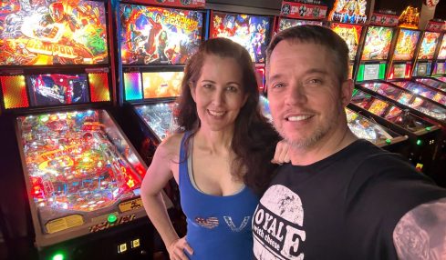 A smiling man and woman take a selfie in front of pinball machines at an arcade.