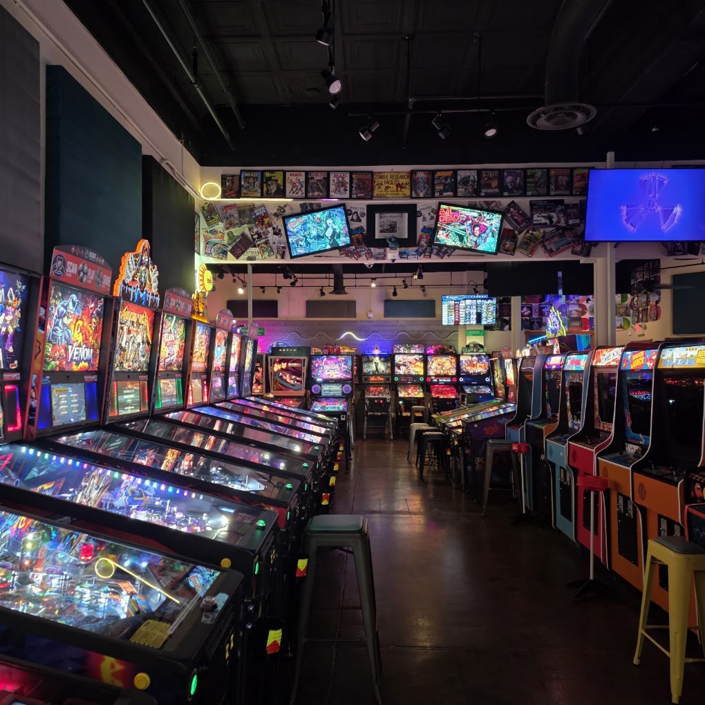 Arcade with rows of colorful pinball machines and video game screens, dim lighting, and seating area, lively atmosphere.