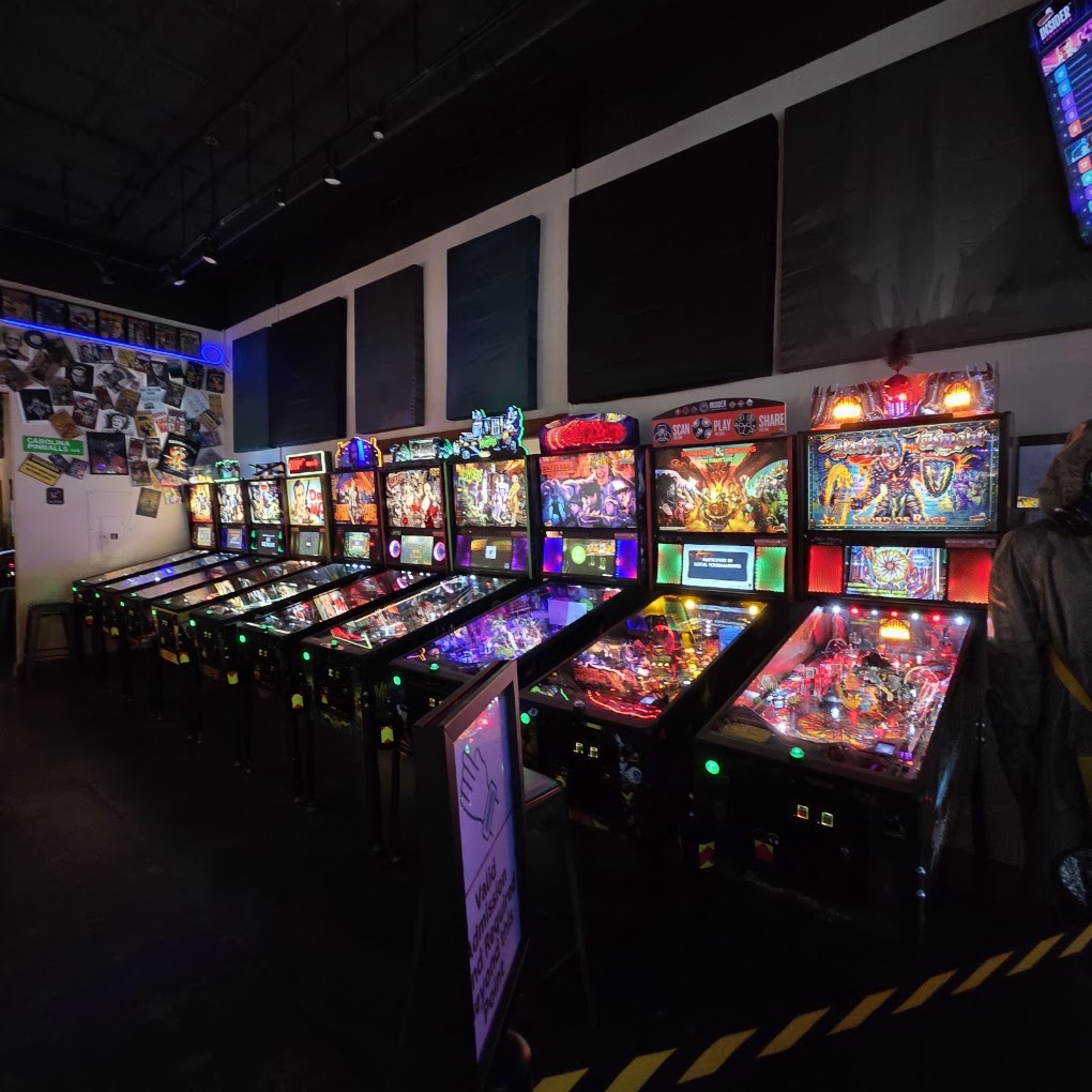 Row of pinball machines in a dimly lit arcade with colorful lights and a wall with photos and blackboards.