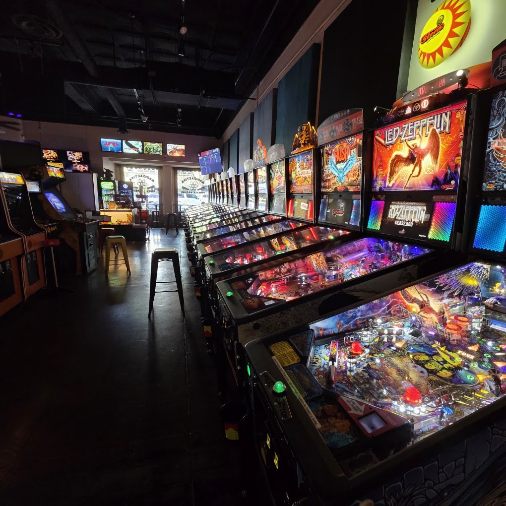 Arcade with pinball machines and gaming screens, dimly lit with colorful lights, and a seating area in the background.