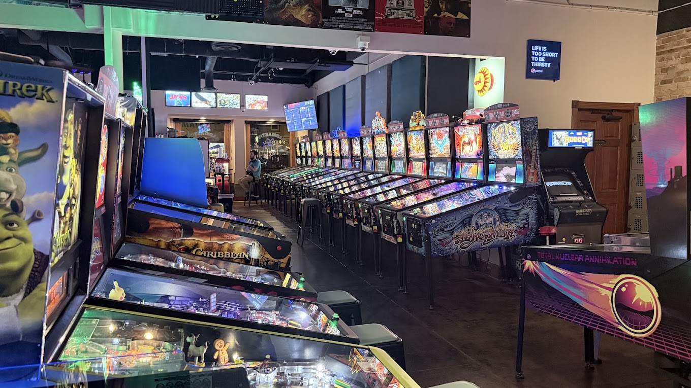 Arcade with pinball machines and colorful video game screens, illuminated by bright lights, in a gaming center.