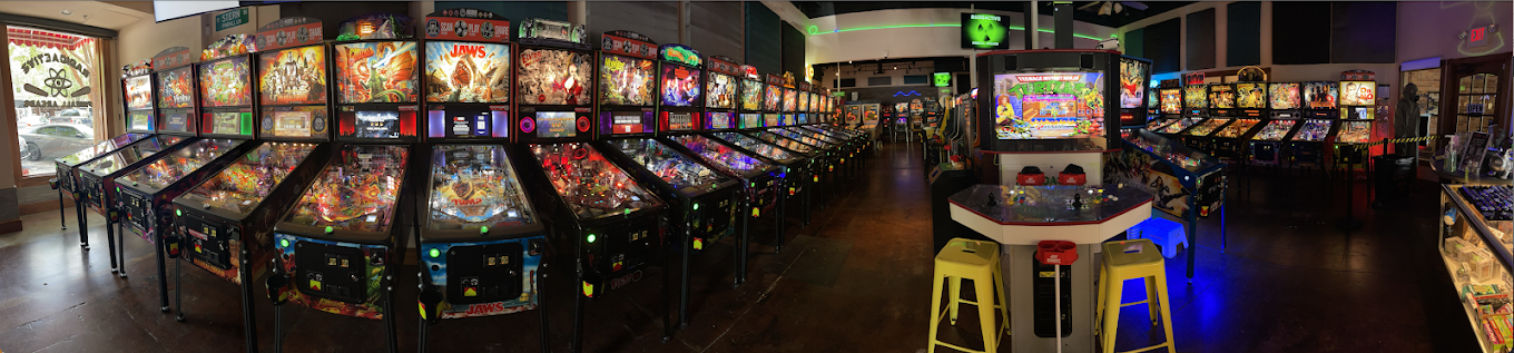 Arcade with multiple pinball machines and colorful screens, seating area with yellow and blue stools, dim lighting.