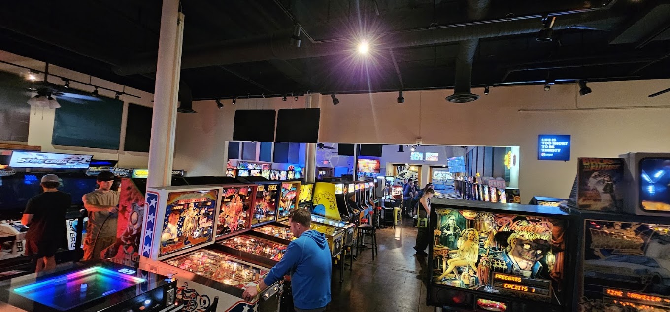 Arcade with pinball machines and people playing games, illuminated by bright lights, in a lively indoor setting.