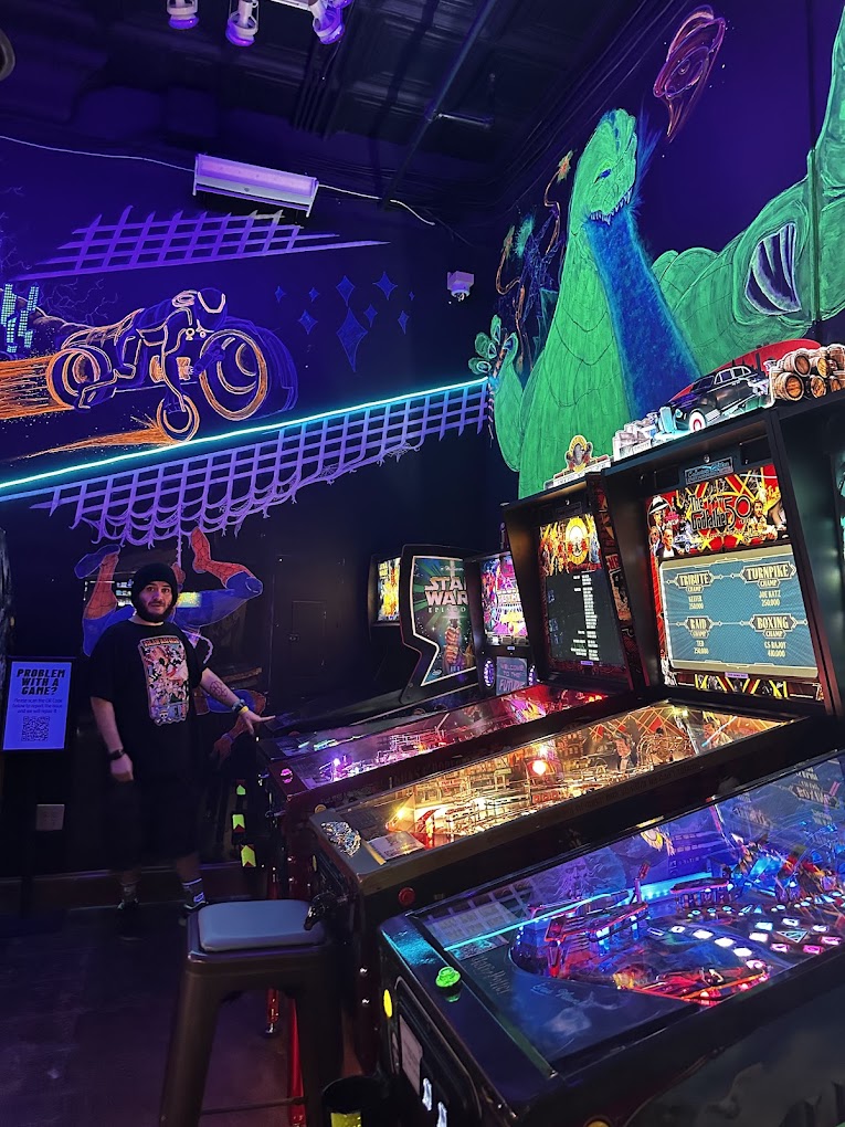 Arcade with pinball machines, neon lights, and a large colorful dinosaur mural on the wall, with two people standing nearby.