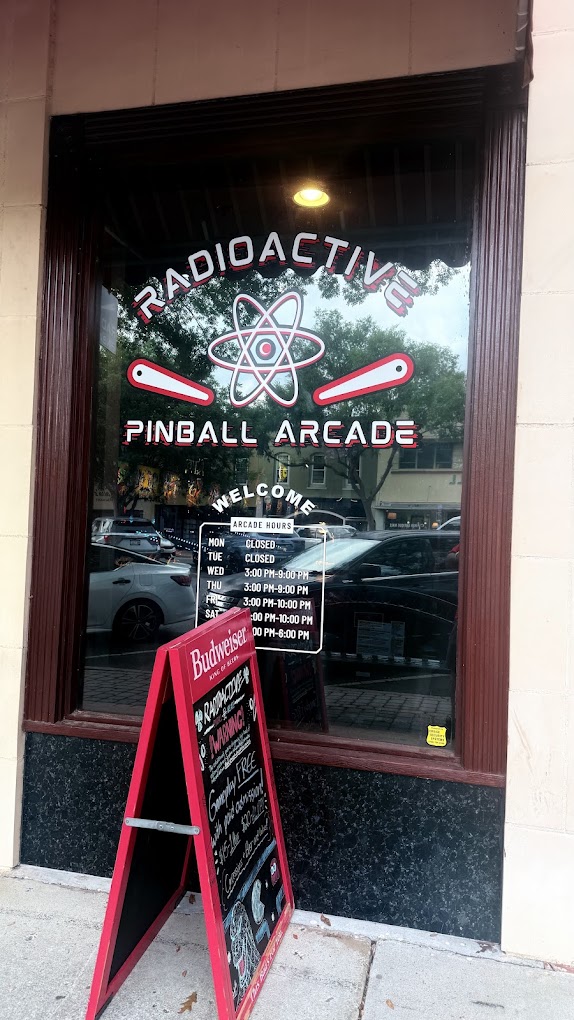 Window with radioactivity symbol, text for radioactiv Pinball Arcade, and a menu board outside, with cars reflected in the glass.