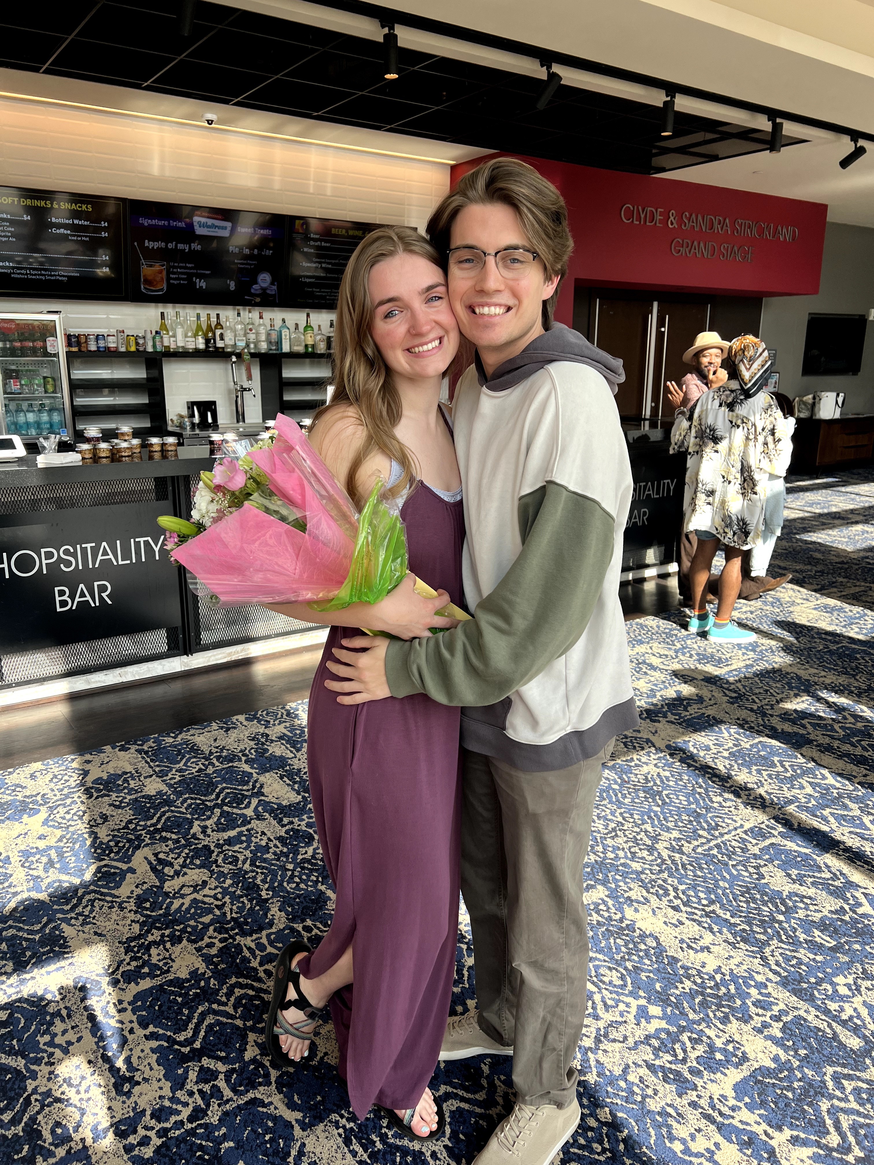 Two people hugging, smiling, in an indoor setting with a bar and other people in the background.