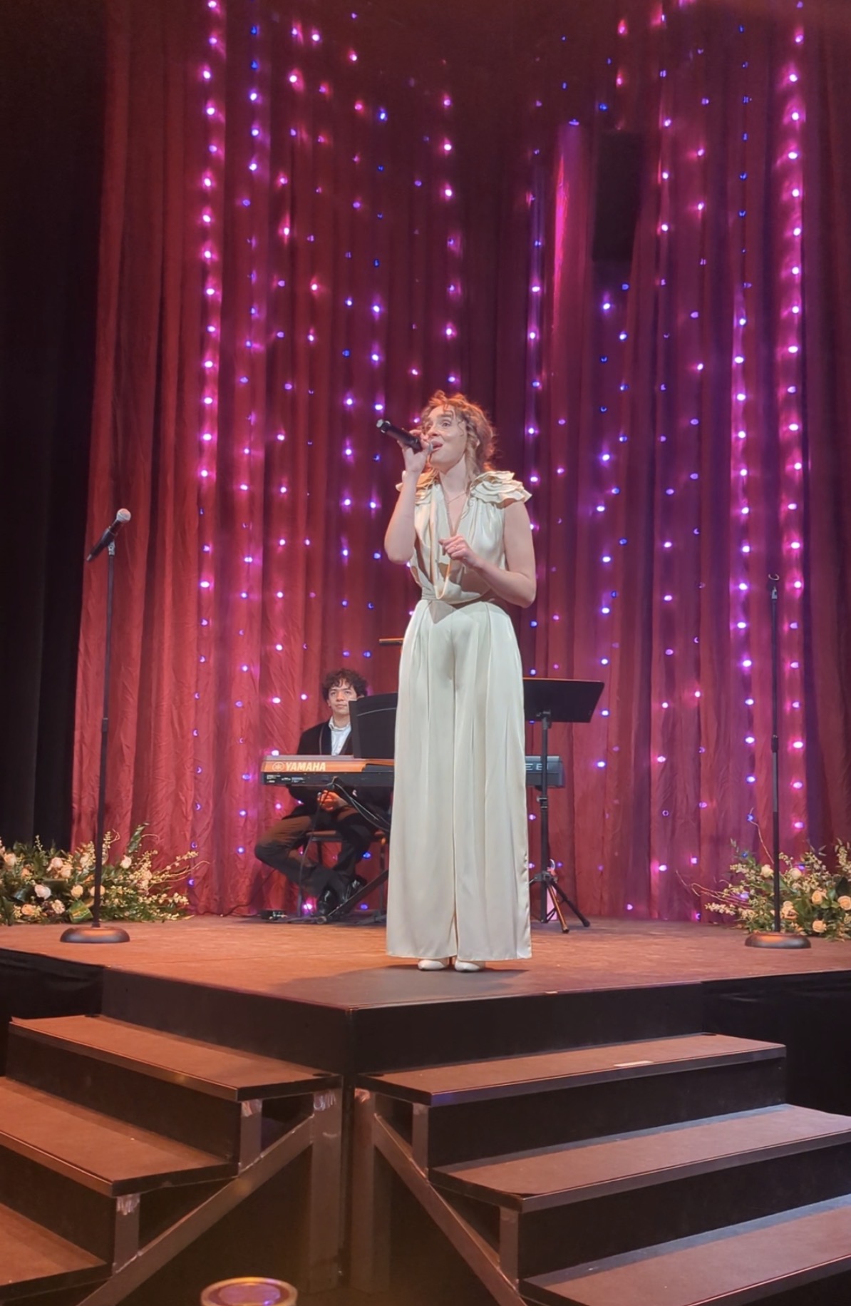 A woman in a white dress sings on stage with pink and purple lights, a keyboard player, and floral decorations.