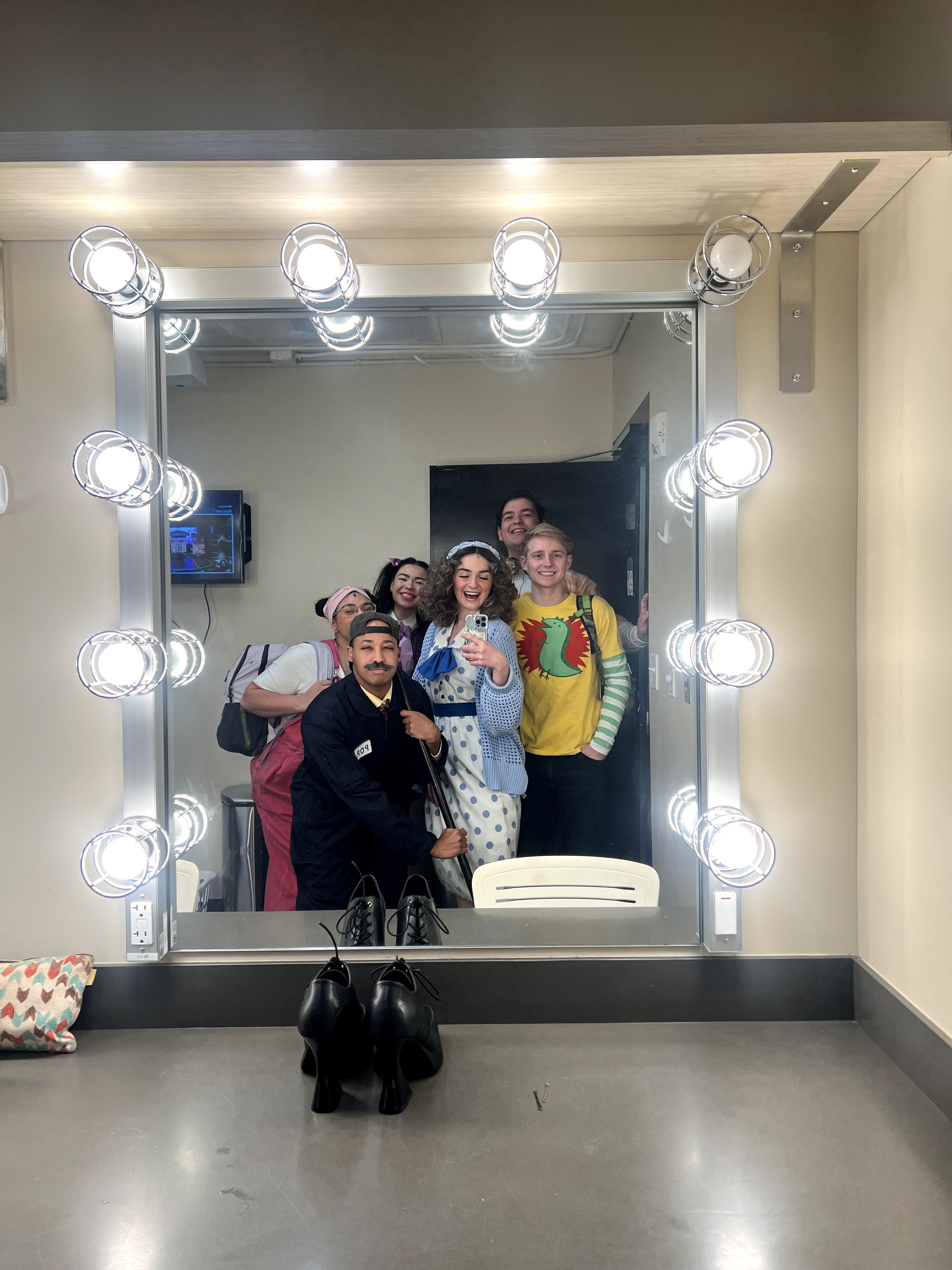 Four people taking a selfie in front of a mirror with bright lights, in a dressing room or backstage area.
