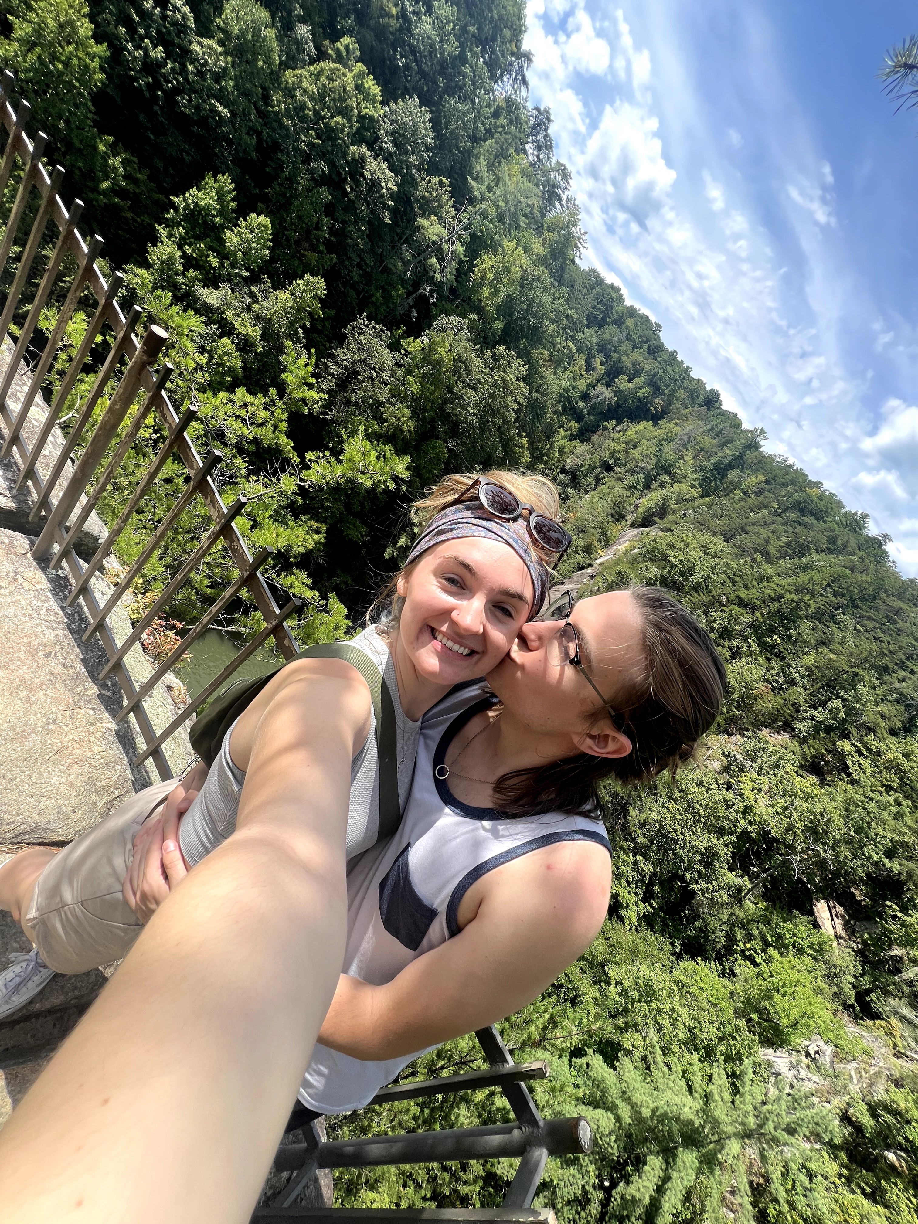 Two women smiling and hugging outdoors with trees and a blue sky in the background.