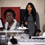 A woman in a pinstripe suit stands behind a table with jewelry, with a large portrait of another woman in the background.