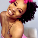Smiling woman with pink and black curly hair, wearing a pink top and necklace, showing teeth and closed eyes.