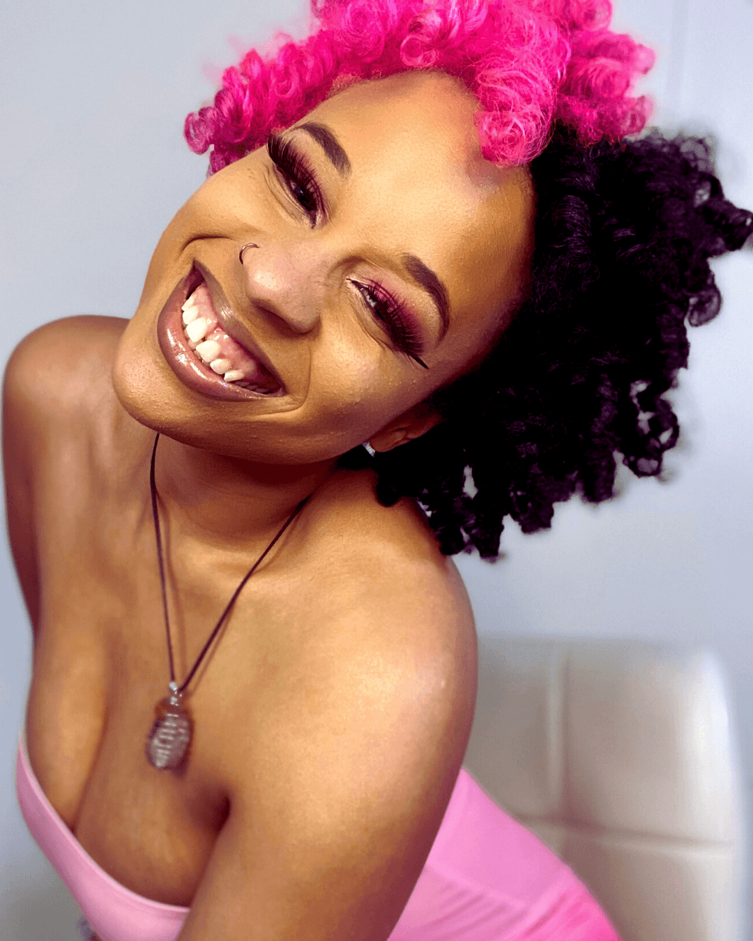 Smiling woman with pink and black curly hair, wearing a pink top and necklace, showing teeth and closed eyes.