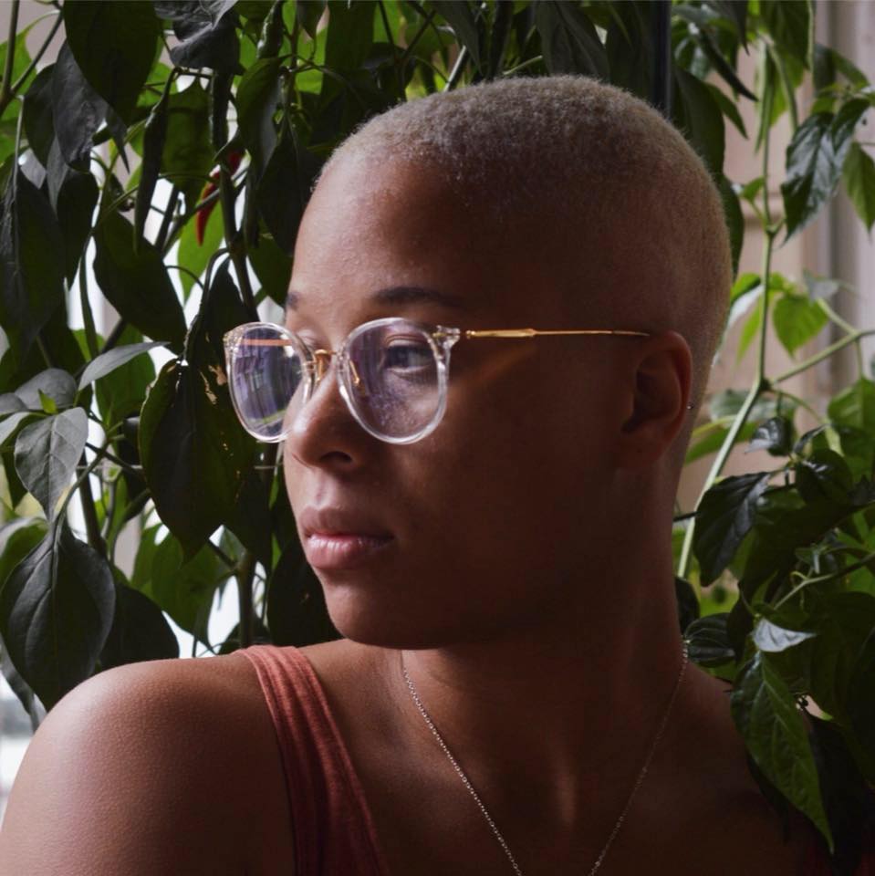 Young person with short hair and glasses looking to the side among green plants.