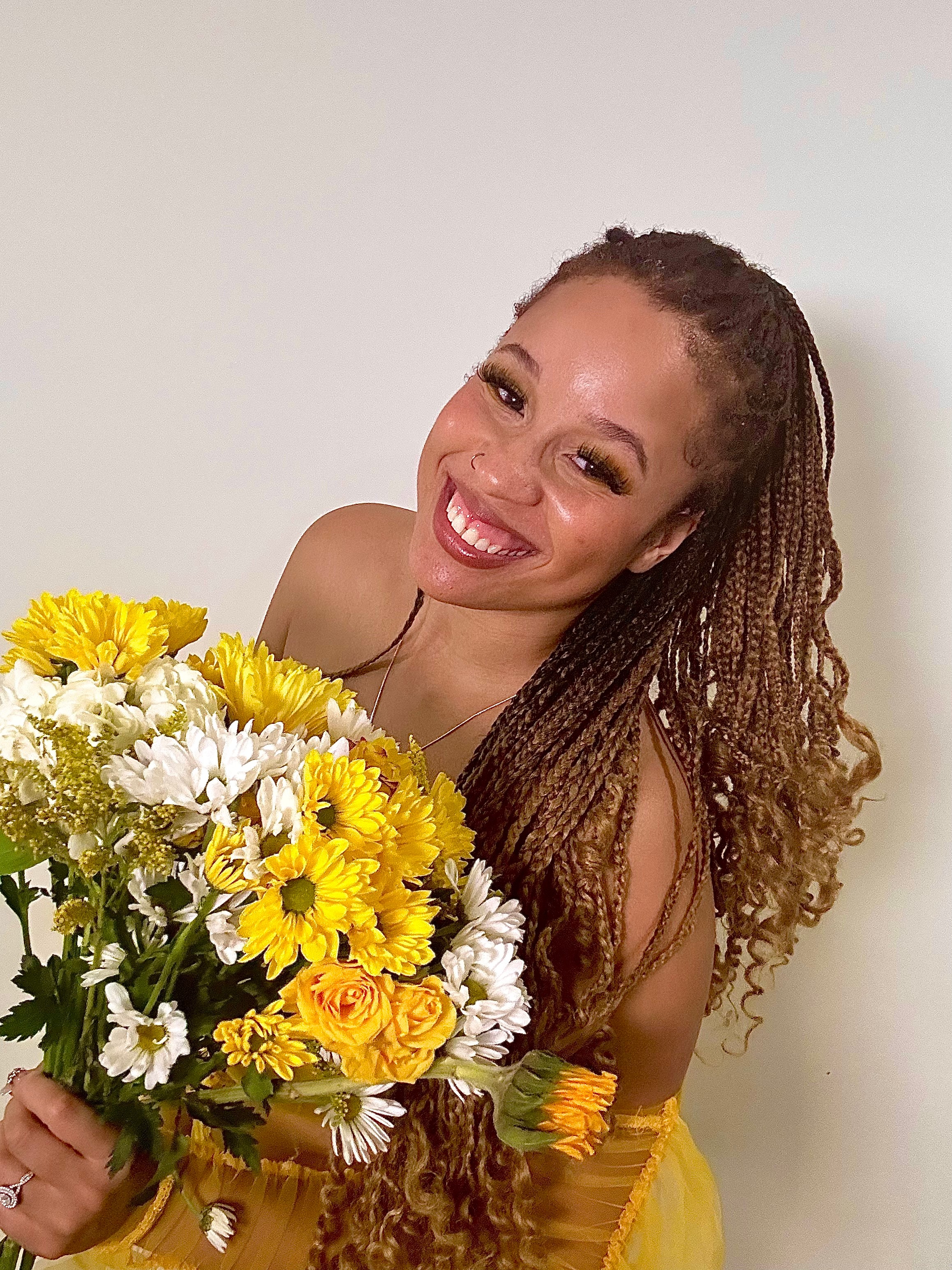 Smiling woman with long braided hair holding a large bouquet of yellow and white flowers, wearing a yellow top.
