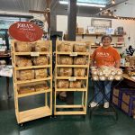 Person standing behind a wooden display shelf with bread in a store, with a red sign reading 'Toys' nearby.