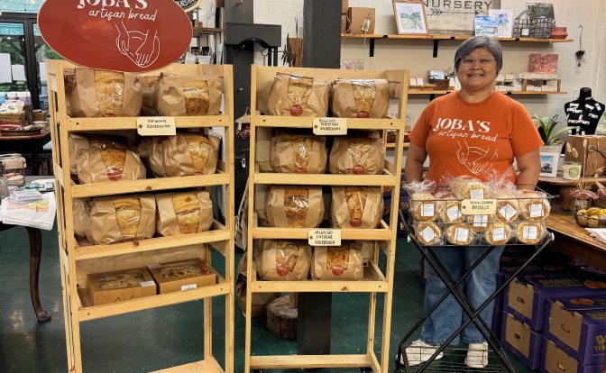 Person standing behind a wooden display shelf with bread in a store, with a red sign reading 'Toys' nearby.