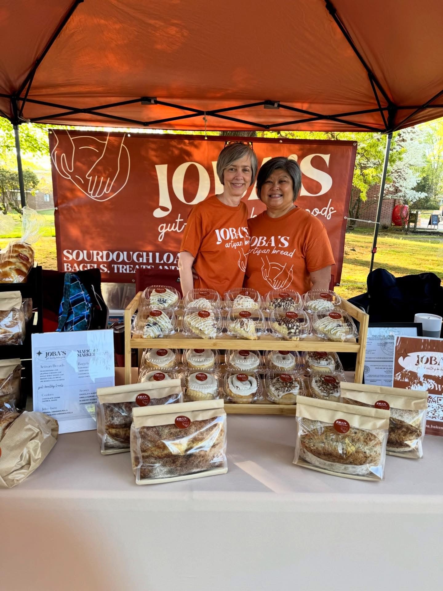 Two women stand behind a table with baked goods at an outdoor market stall, smiling, with a red banner in the background.