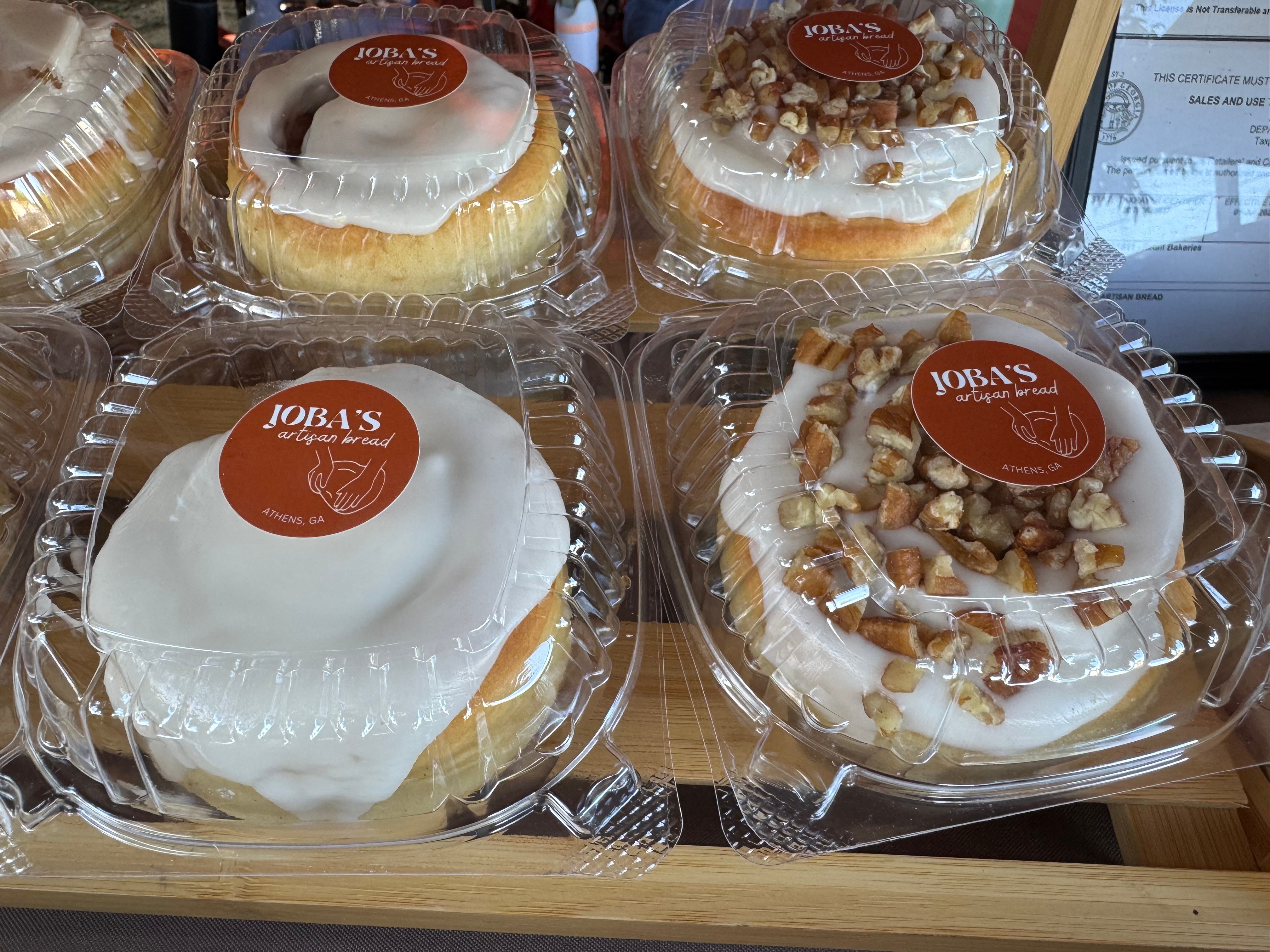Four cakes in plastic containers, two with white icing and two with nuts and caramel topping, on a wooden surface.