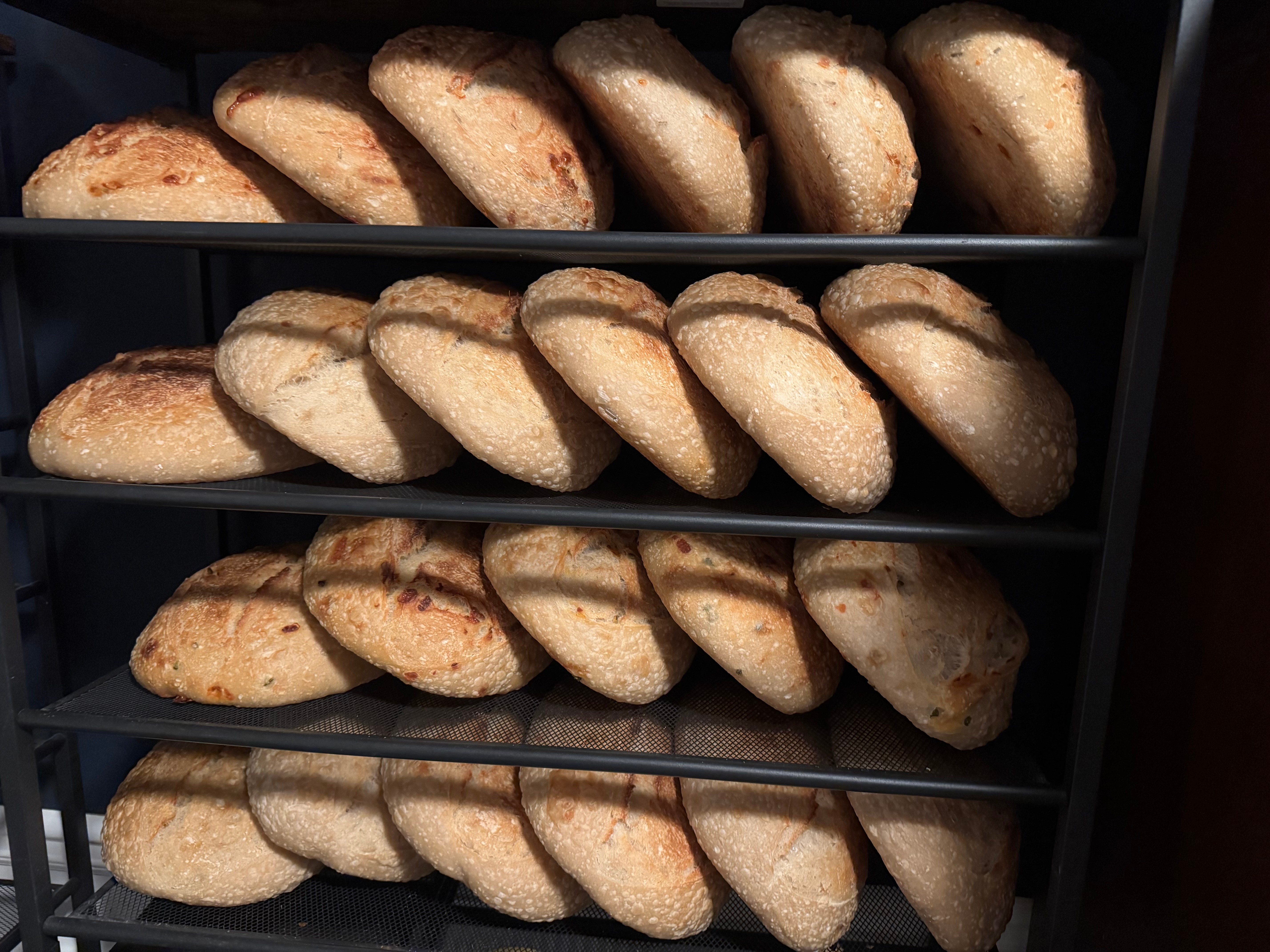 Multiple loaves of bread on a black metal rack, arranged on four shelves, with a variety of shapes and sizes.