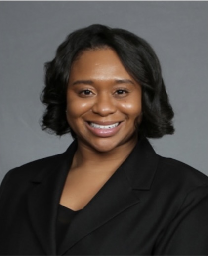 Portrait of a smiling woman with shoulder-length wavy hair, wearing a black blazer against a gray background.