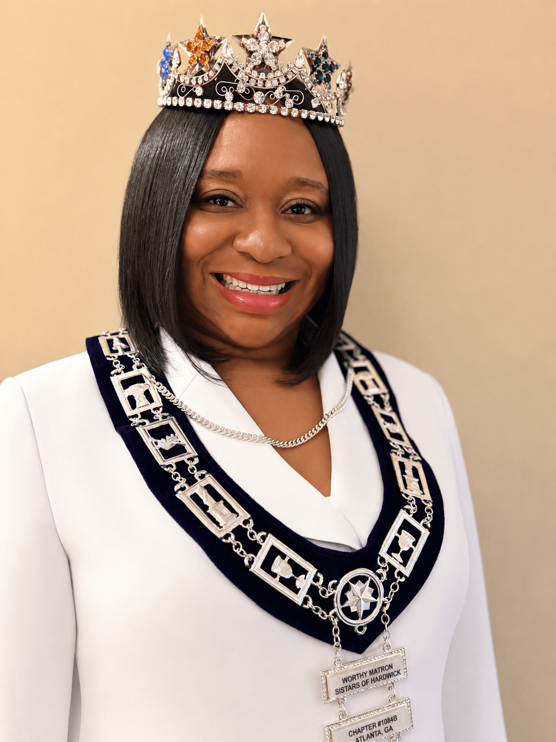 Woman wearing a crown and a white blazer with a decorative chain, smiling, against a plain background.