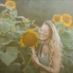 Woman with glasses smiling in a sunflower field, holding a sunflower close to her face, surrounded by tall sunflowers.