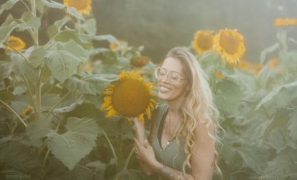 Woman with glasses smiling in a sunflower field, holding a sunflower close to her face, surrounded by tall sunflowers.