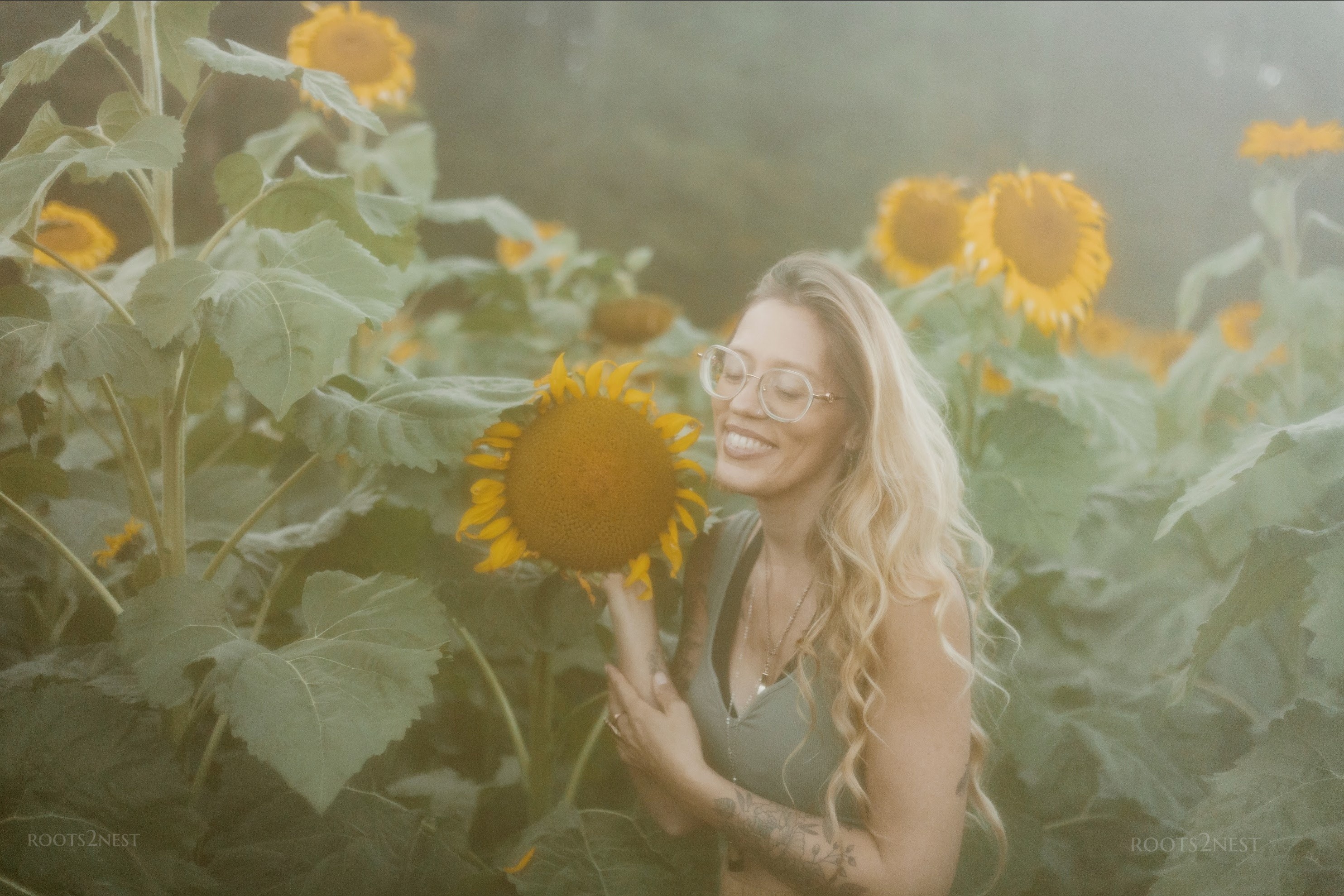 Woman with glasses smiling in a sunflower field, holding a sunflower close to her face, surrounded by tall sunflowers.