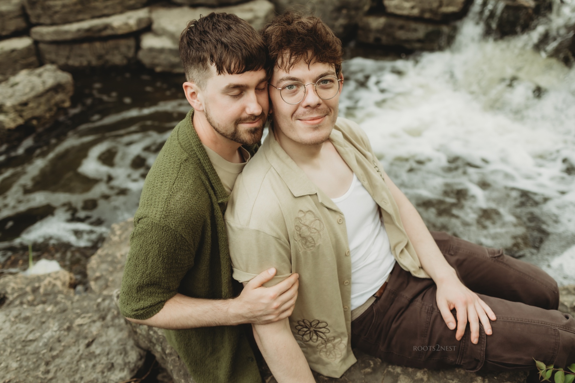 Two young men sitting on rocks near a flowing stream, smiling and embracing each other outdoors.
