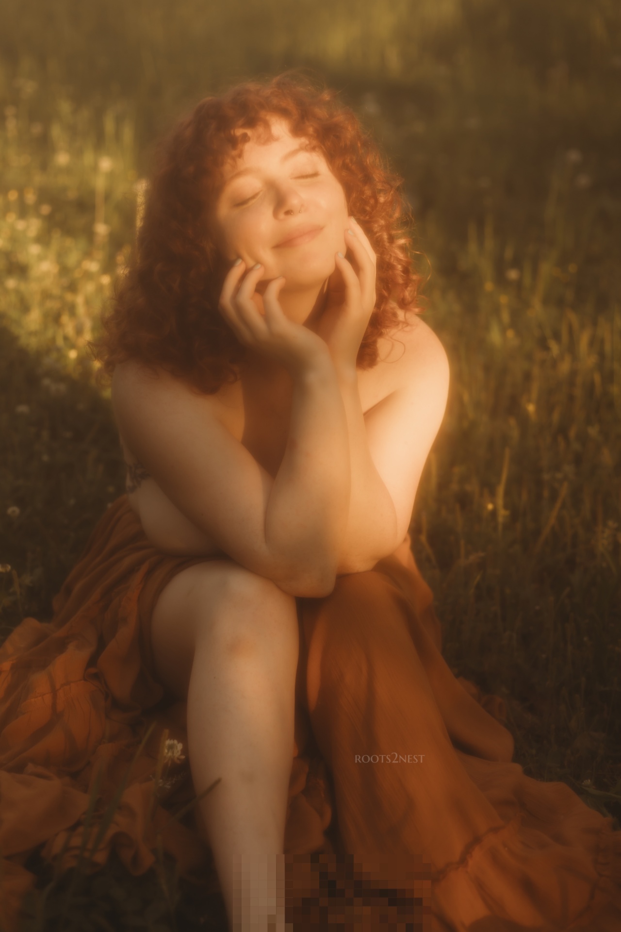Young woman with curly hair sitting outdoors, smiling with eyes closed, hands on cheeks, wearing a brown dress.