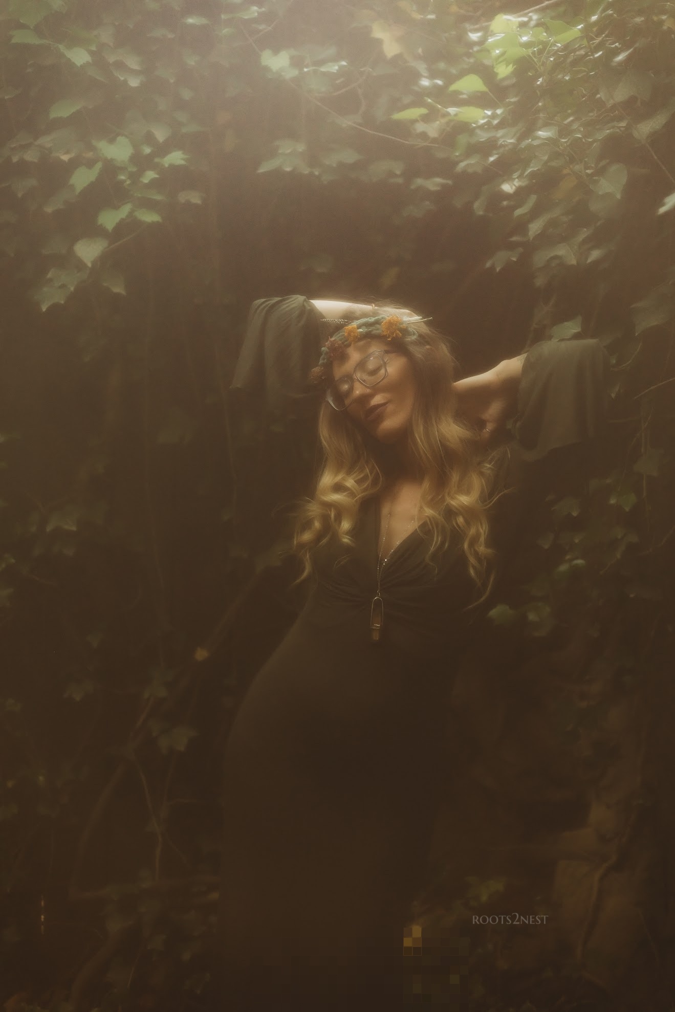 Woman with long curly hair in a dark forest, arms raised, surrounded by dense foliage.