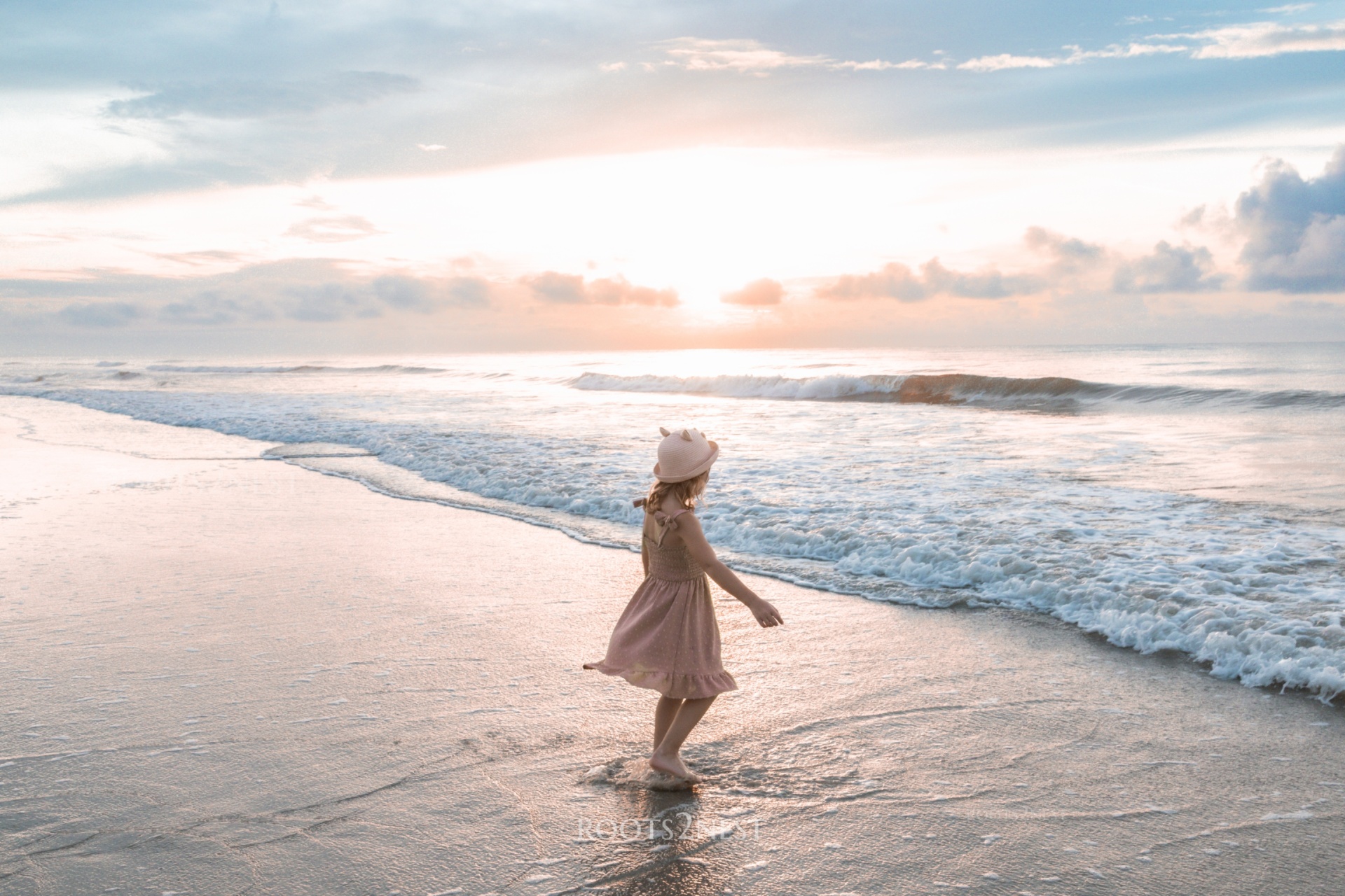A girl in a pink dress and hat standing in shallow ocean water at sunset, with waves and sky in background.