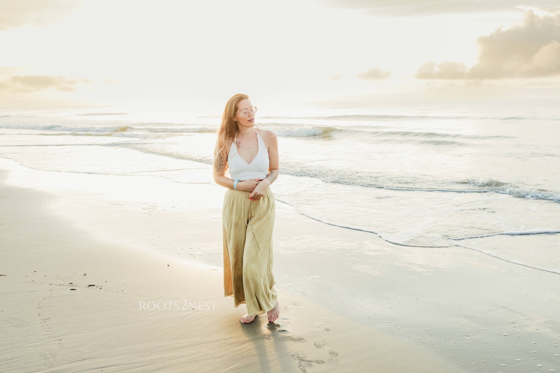 Woman walking on a beach at sunset, wearing a white top and long skirt, looking to the side.