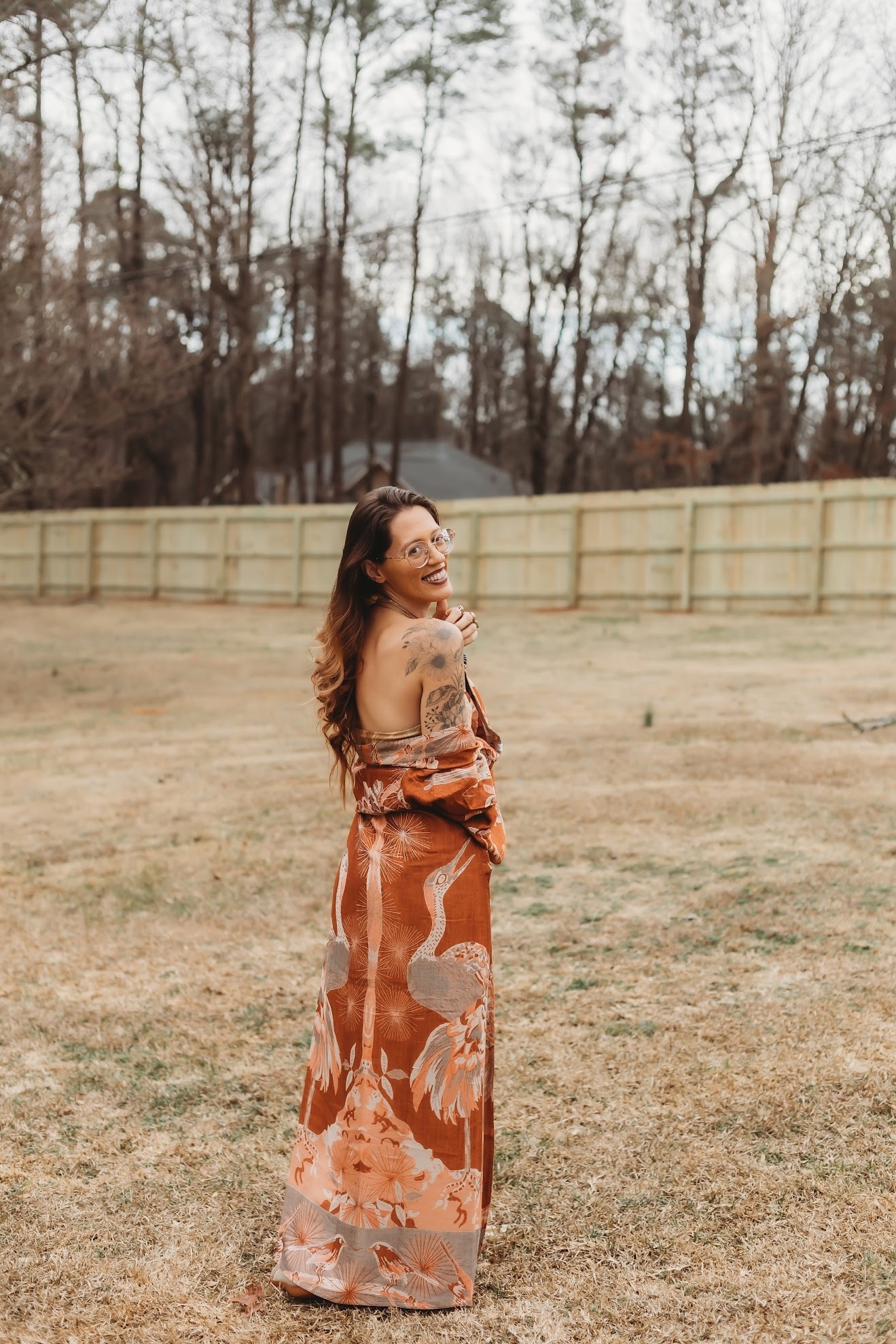 Woman in a long dress standing outdoors on grass, smiling, with trees and a fence in the background.