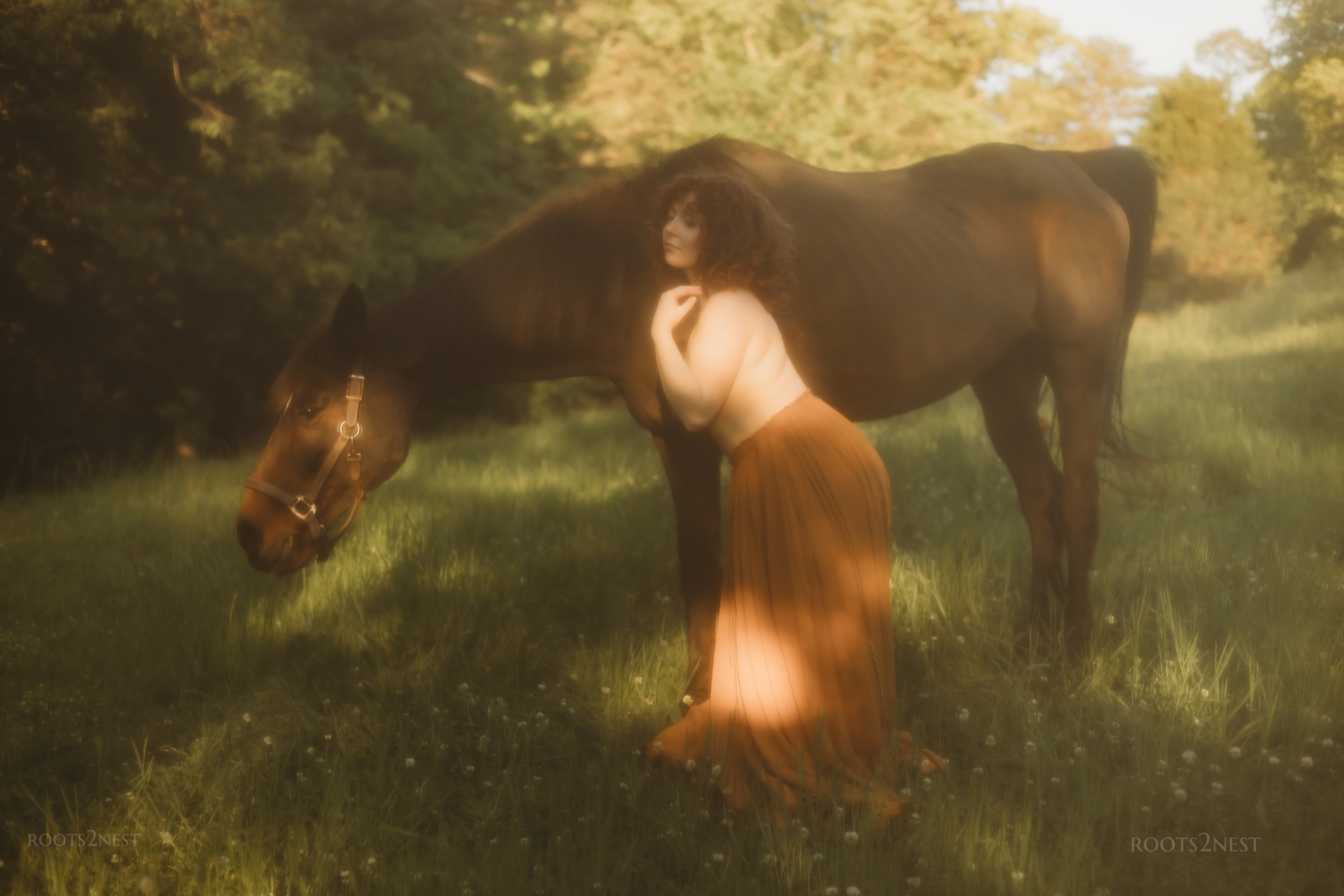 Person bending over a horse in a grassy field with trees in the background.
