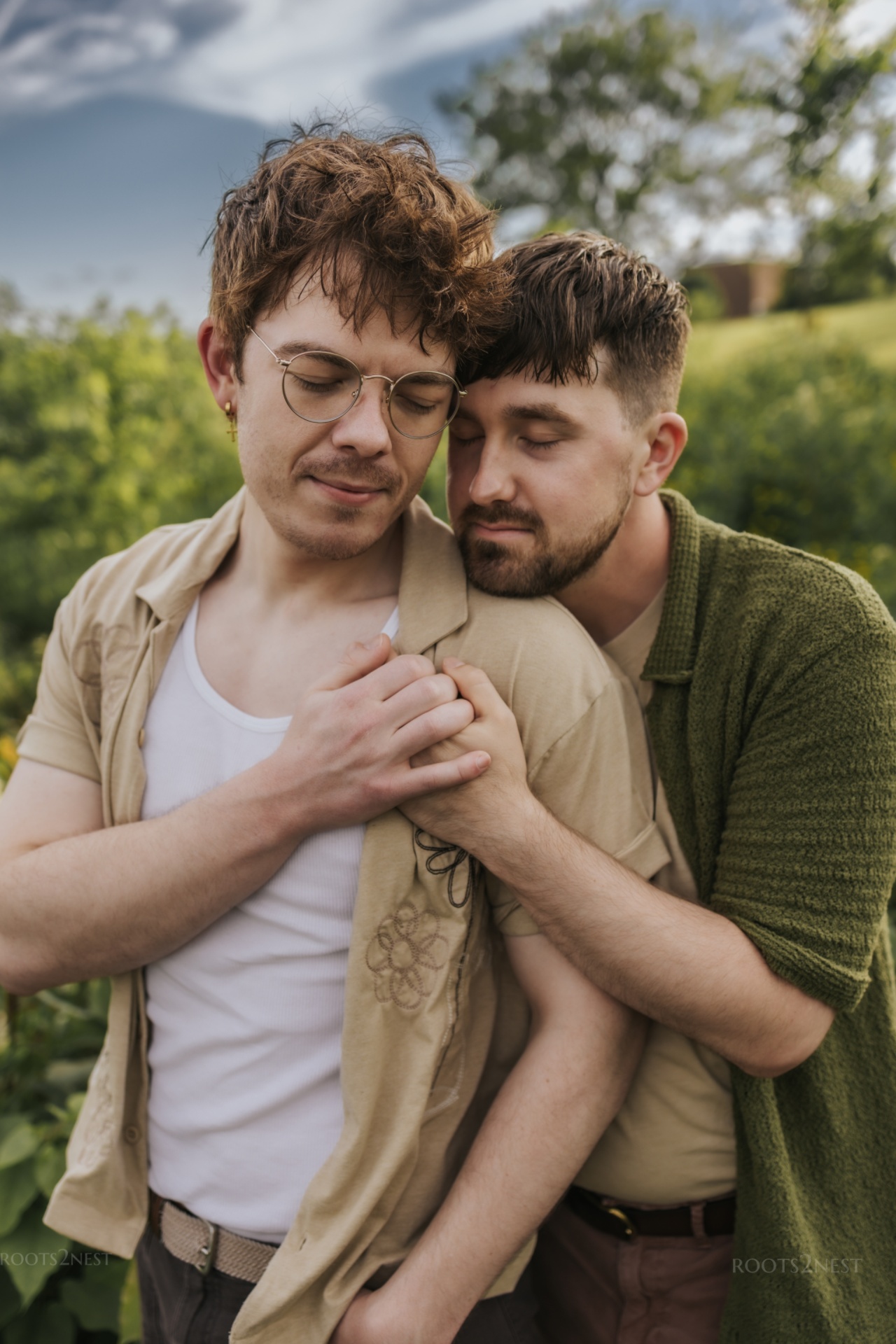 Two men embrace outdoors, one with glasses and curly hair, the other with short dark hair, both with eyes closed.